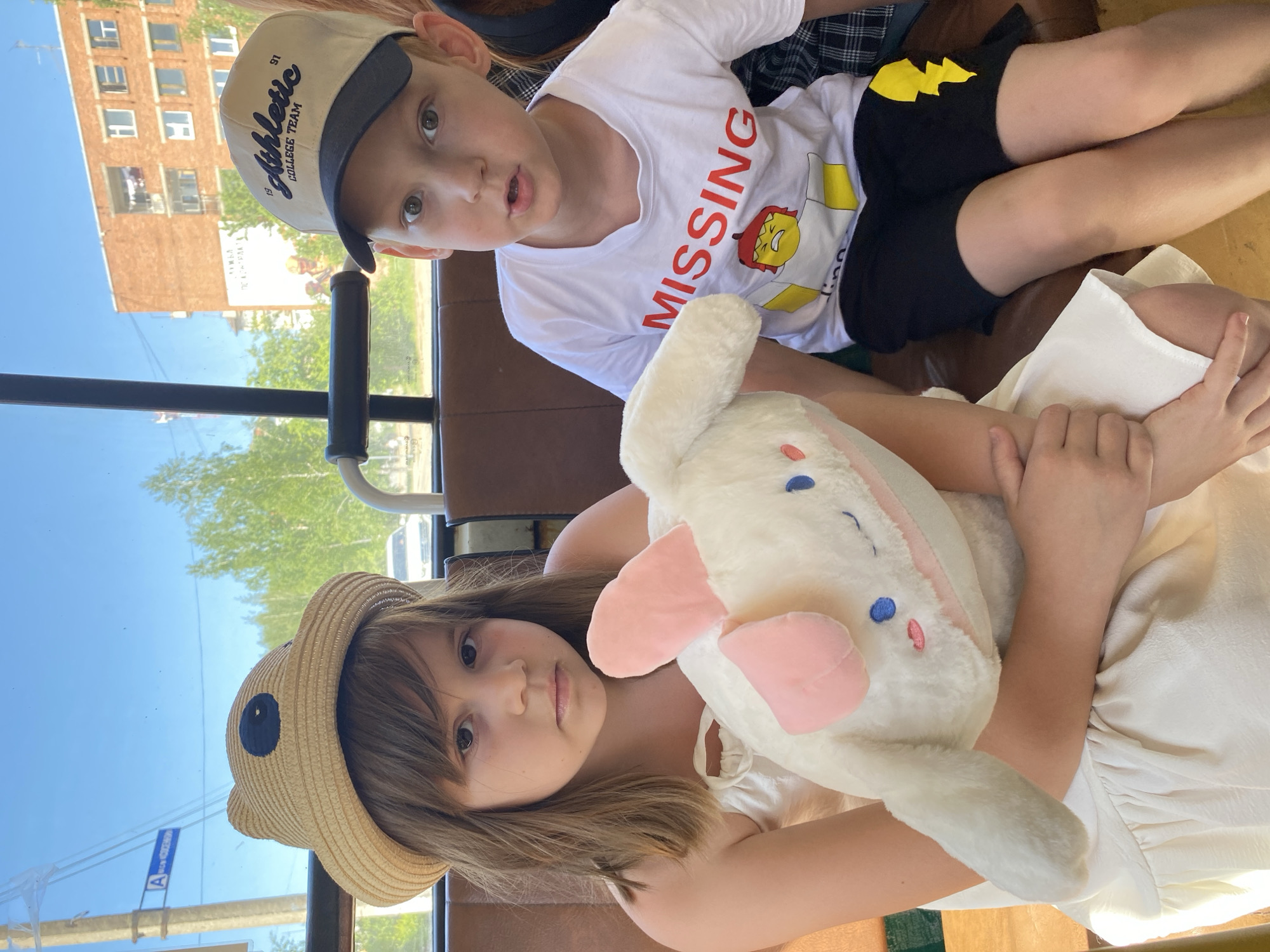 Two children are seated inside a bus or similar public transportation vehicle. The younger child, a girl with light brown hair and wearing a straw hat with bear ear appliques, is sitting in the foreground and holding a large, stuffed white rabbit toy with pink ears. The older child, a boy wearing a baseball cap and a white t-shirt with the word "MISSING" in red, sits behind her. The background through the window shows buildings and trees, indicating an urban setting. A street sign in Cyrillic script is visible through the window. The location appears to be Noviy, Russia.