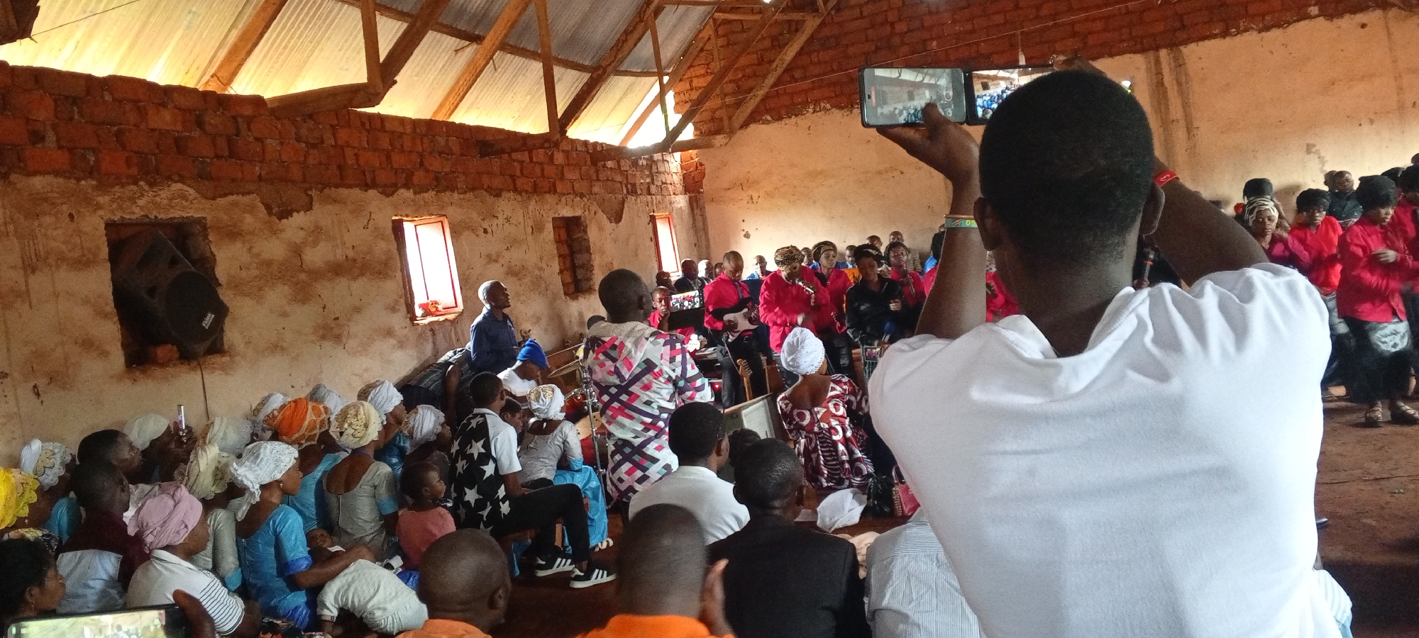 In Ngalula, Democratic Republic of the Congo, an indoor gathering takes place. An individual in the foreground, seen from behind and wearing a white shirt, holds up a mobile phone as if recording the event. The room appears to be a place of worship or community meeting space, with numerous people present. Many individuals on the left side of the frame wear matching light blue outfits and white head coverings. A group of people in the background are dressed in red attire, possibly singing or performing. The structure's interior has bare brick walls and a roof supported by wooden beams. One brick-framed opening in the wall houses a dark speaker. Several windows provide natural light.