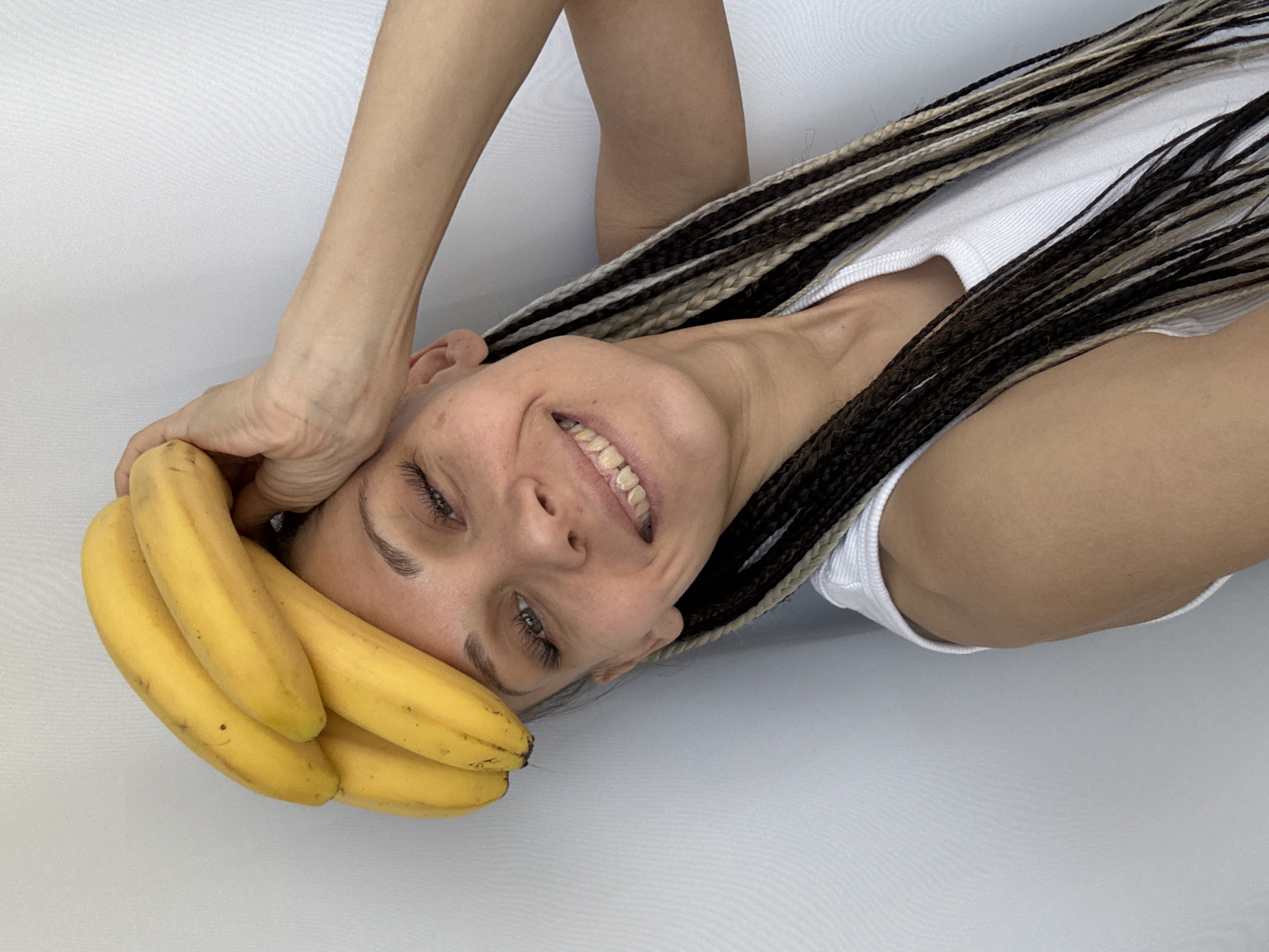 A smiling woman is holding a bunch of bananas to her forehead. She has braided hair in dark brown and blonde shades. The background is plain white. There is no indication of the location being Rusanovo, Russia.