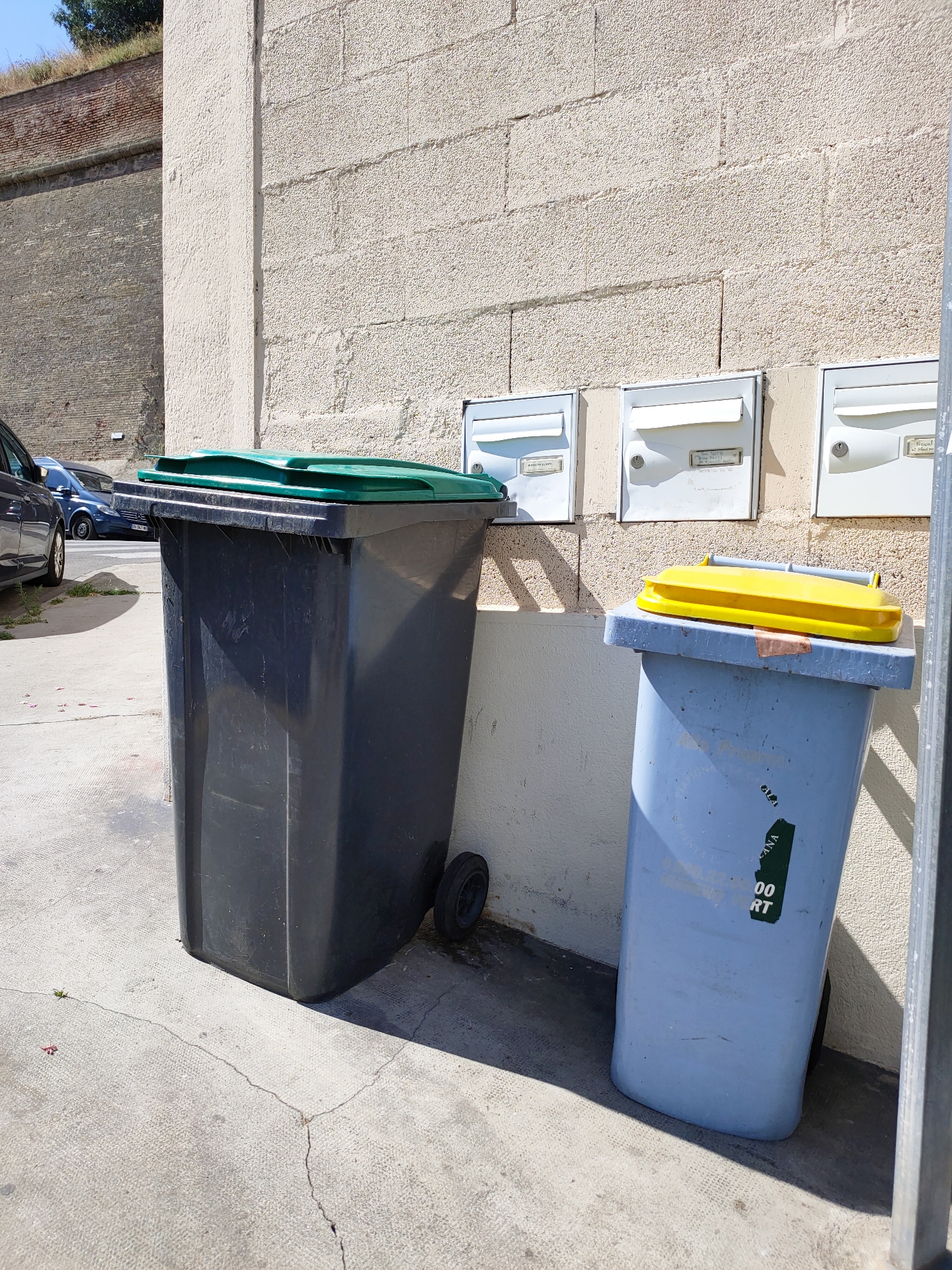 This image depicts a scene outdoors, likely on a sunny day, with a textured, light-colored brick wall forming the background. In the foreground, two large garbage bins are prominent. The bin on the left is dark gray with a green lid, and it has wheels on the bottom. To its right is a light blue bin with a bright yellow lid. A sticker with text is visible on the side of the blue bin, partially obscured, but "00 RT" is legible. Above the bins, mounted on the wall, are three white mailboxes with their flaps down. The mailbox on the far right has some writing visible, possibly a name or address.
The ground is concrete, with visible cracks and some sparse green shoots. To the left of the garbage bins, a portion of a dark-colored car is visible, suggesting a parking area or street. In the upper left corner of the image, a glimpse of a brick wall, possibly a historical fortification or embankment, topped with dry grass, is visible, hinting at the broader location. The sunlight casts distinct shadows, indicating it is likely midday. The overall impression is one of an everyday urban or semi-urban setting. No people are visible in the frame. The mood is neutral and observational.