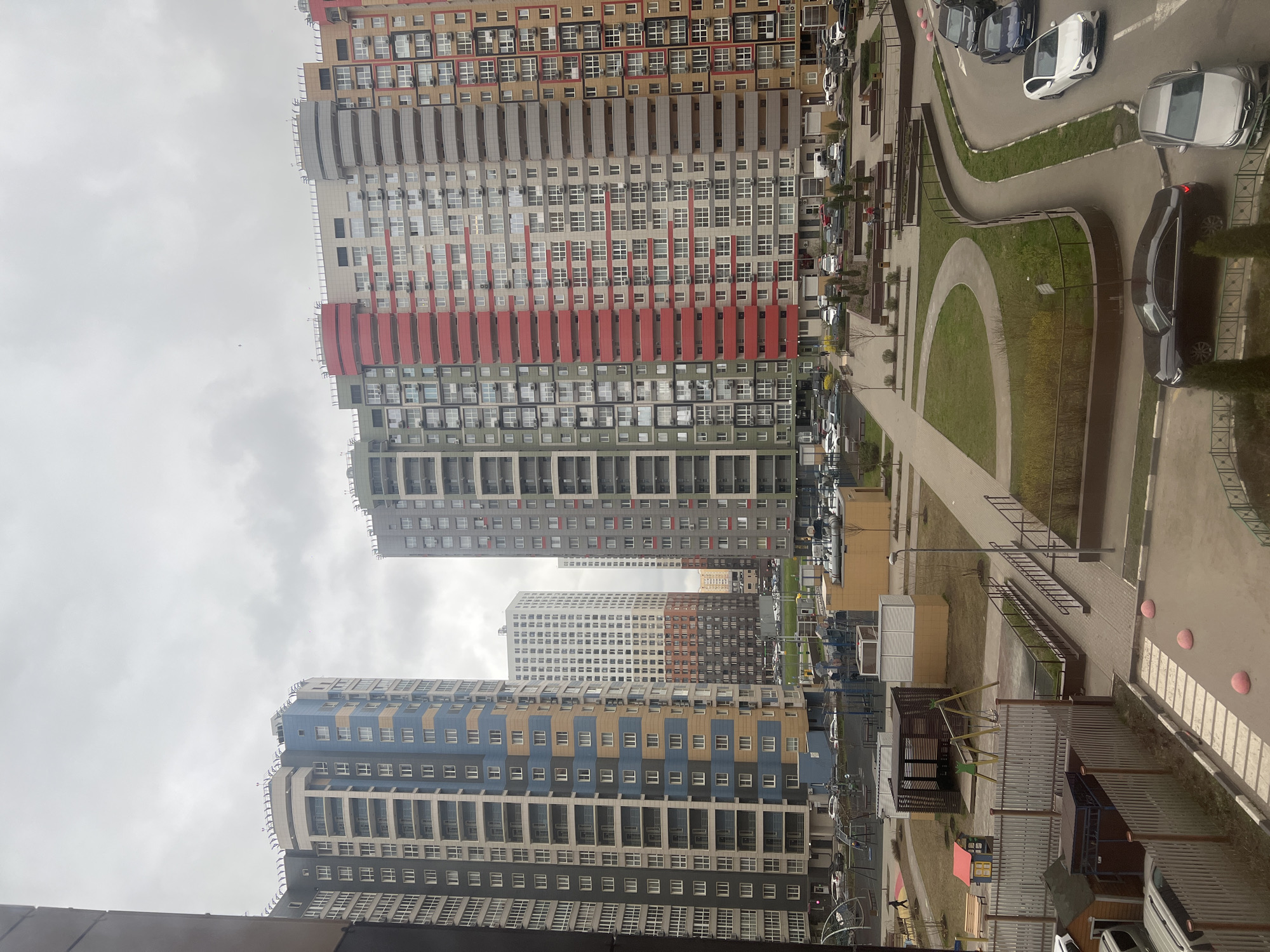 The image depicts a view of residential buildings in Khimki, Russia. The buildings are tall, multi-storied structures with a variety of colors on the facades, including blue, grey, red, and beige. Each building has a multitude of windows, indicating apartment units.
Between the buildings, a courtyard area is visible, featuring walkways, greenery, parking spaces with cars, and a playground. The sky is overcast, suggesting a cloudy day. The overall scene conveys a densely populated urban environment.