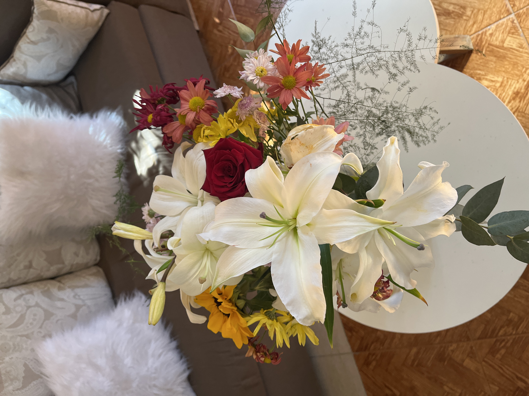 The image shows a floral arrangement on a white, two-tiered, oval-shaped table. The arrangement features large white lilies, a red rose, and various smaller flowers in shades of orange, yellow, and pink. Some greenery is also present. The table is situated on a wooden floor with a parquet pattern. In the background, part of a gray couch is visible, adorned with light-colored pillows. The location is San Fernando del Valle de Catamarca, Argentina.