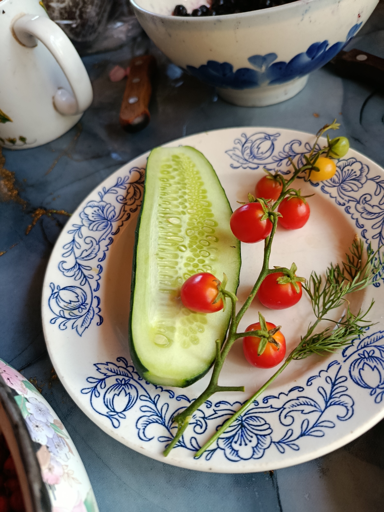 A close-up, overhead shot shows a slice of cucumber and a vine of cherry tomatoes on a white plate with a blue floral pattern. The plate is set on a dark blue surface with gold accents. To the left of the plate is a white pitcher with a gold handle, and behind it, a bowl of dark berries. A small knife with a wooden handle lies between the pitcher and the bowl. In the background, another bowl with a blue and white floral pattern is visible, containing what appears to be berries. The scene suggests a meal preparation or serving setting, possibly in a home environment within the city of Vereya, Russia.