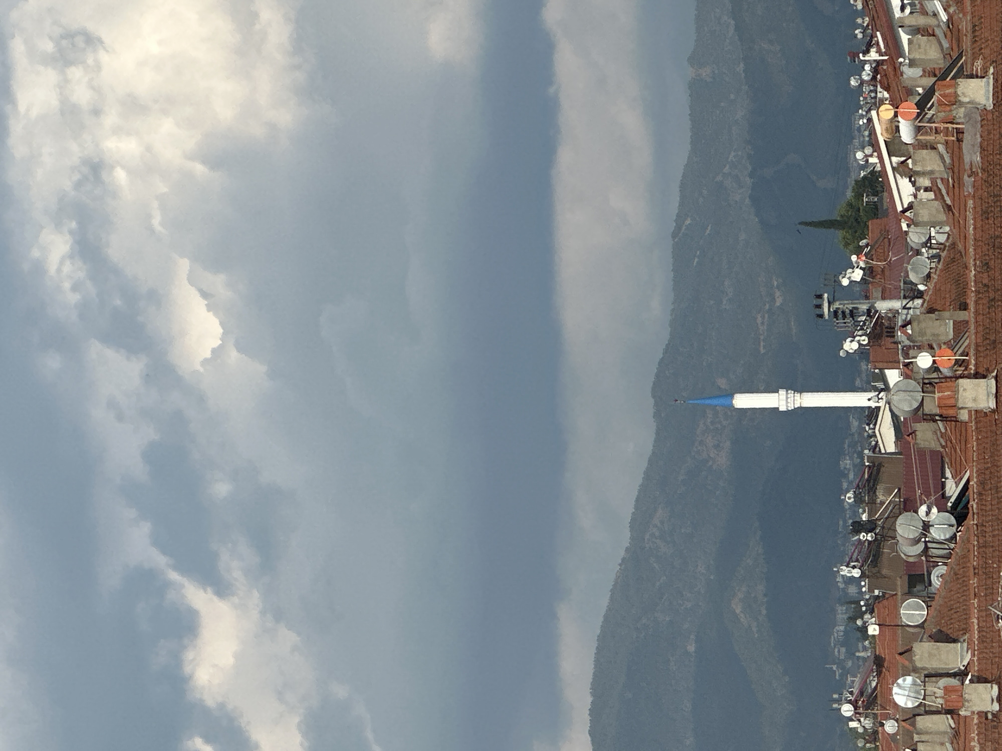 The image shows a view from an elevated position in Fethiye, Turkey. The right of the image depicts a row of terracotta tiled roofs, heavily populated with satellite dishes and solar water heaters. Beyond the rooftops lies a mountainous region, partly obscured by haze. A slender, white minaret with a blue conical roof stands out against the mountain backdrop. To the left, the sky is filled with dramatic, overcast clouds.