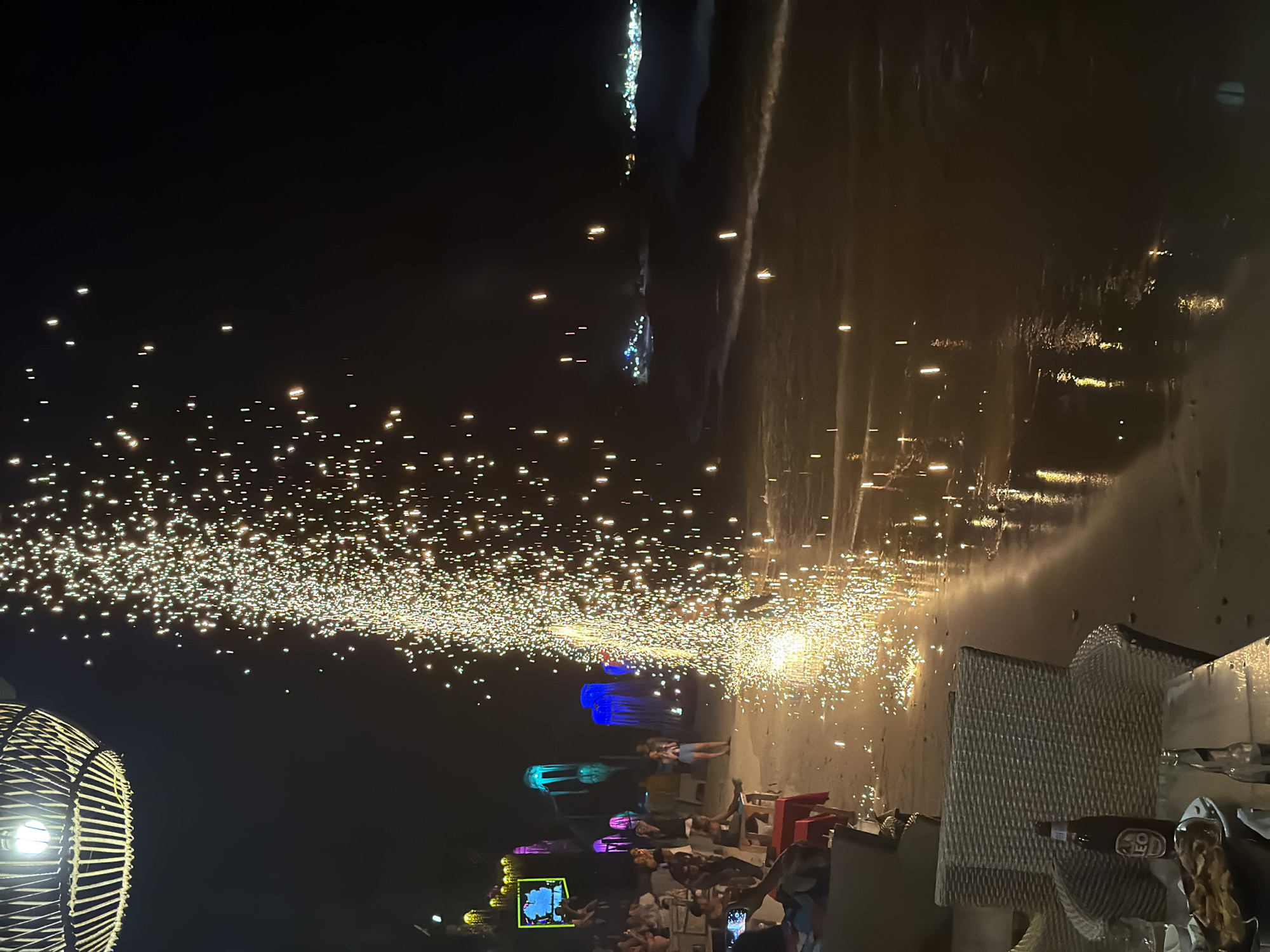 The image depicts a nighttime scene on a beach in Ao Phai, Thailand. A large, ground-based pyrotechnic device, emitting a dense vertical spray of golden sparks, is active near the waterline in the mid-ground, illuminating the wet sand and casting reflections. To the left of the pyrotechnic display, multiple individuals are gathered on the sand. Some are seated at low tables with chairs, while others stand observing the display. In the immediate foreground, an overhead wicker lantern with a single bulb is visible in the upper left corner, casting light downward. Below it, a table holds a brown bottle identified as "LEO" beer, another glass bottle, and a plate containing food items resembling chips or nachos. A wicker chair is positioned to the right of this table.
Further into the background on the beach, a series of illuminated decorative structures resembling glowing jellyfish or fantasy plants in blue, pink, green, and yellow hues are visible. Other groups of people are scattered across the beach area. In the far background, across the water, distant clusters of artificial lights indicate a shoreline or vessels. The ocean shoreline shows gentle waves breaking, with light reflecting off the wet sand and water. The overall environment suggests an evening gathering or event at a beachside establishment.
