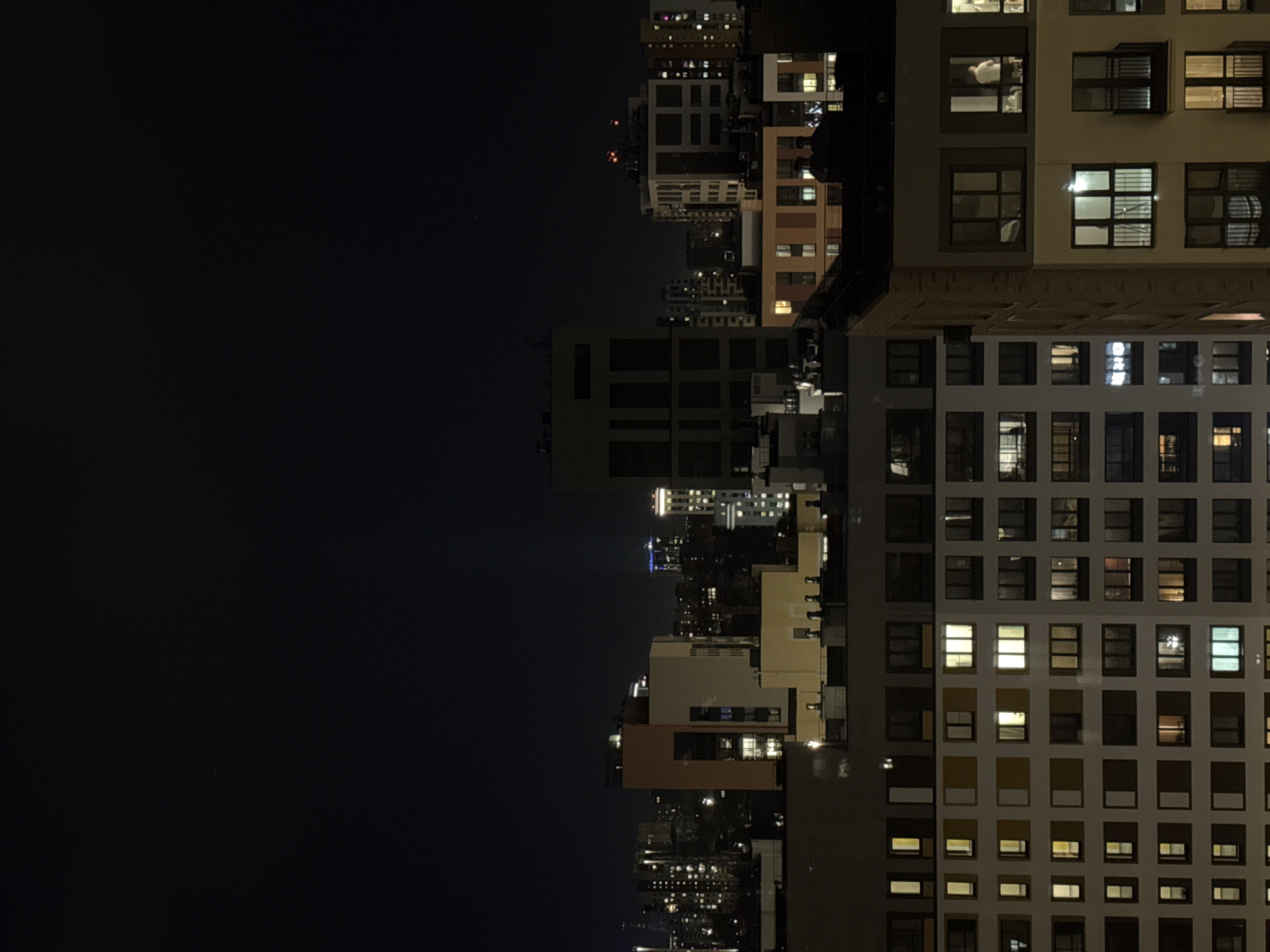 The image is a nighttime view of a city skyline. A large, multi-story building with many windows is positioned on the right side of the frame, with some windows illuminated. To the left, a mix of buildings of varying heights and styles stretch into the distance, and city lights are visible. The left portion of the image is dark, indicating a night sky or shadowed area. The architectural style appears modern, and the lighting suggests it is nighttime in an urban setting. There is no evidence this image takes place in Izhevsk, Russia.
