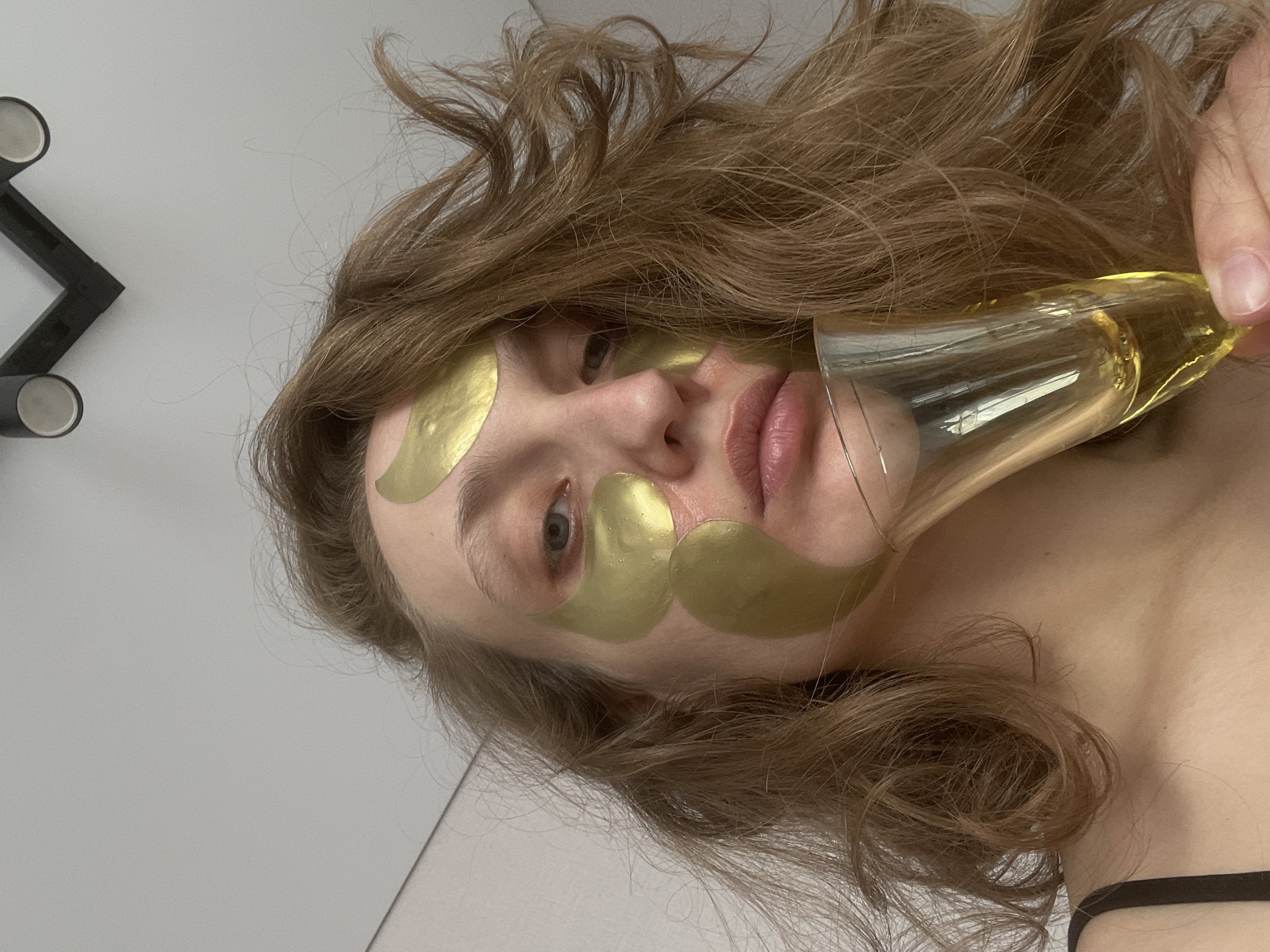The image captures a close-up, sideways view of a young woman engaged in a self-care routine in Подшивалово, Russia. She is lying down, with her head resting on a light-colored surface, and her long, wavy, light brown hair spread around her.
Her face is the central focus, adorned with numerous gold cosmetic patches. Several patches are visible under her eyes, one on her left cheekbone, and multiple others on her forehead and above her left eyebrow. Her eyes are open, looking directly at the viewer, and her lips are slightly parted, revealing a natural pink hue.
In her right hand, which is partially visible on the right side of the frame, she holds a champagne flute filled with a light amber-colored liquid. The glass is held near her face, suggesting she might be about to take a sip or is just holding it as part of her relaxation.
She is wearing a dark-colored, thin-strapped garment, possibly a camisole or bra, visible at the bottom right. The background is a plain, light gray or off-white wall, with a small portion of a black, angular object with two circular, metallic elements visible in the top left corner, possibly a lamp or stand.
The overall scene conveys a sense of calm and relaxation, suggesting a pampering moment. The lighting is soft and diffused, typical of an indoor setting. There is no visible text in the image.