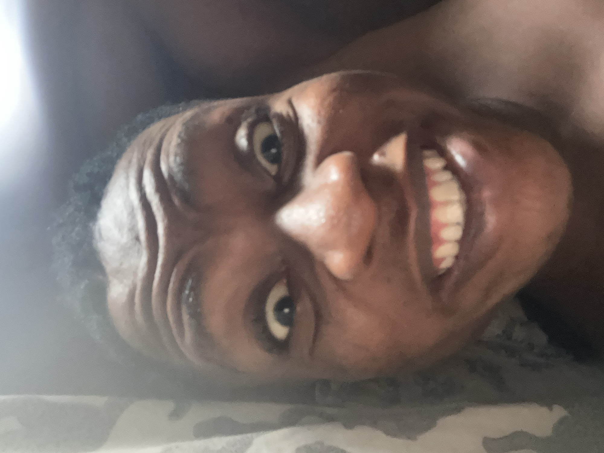The image shows a close-up, rotated view of a person's face. The person has dark skin, visible wrinkles on their forehead, and is smiling, revealing their teeth. The background is out of focus and indistinct, appearing to be bedding with a mottled pattern. A bright light source is present on the left side of the image. The location in the city is unknown.