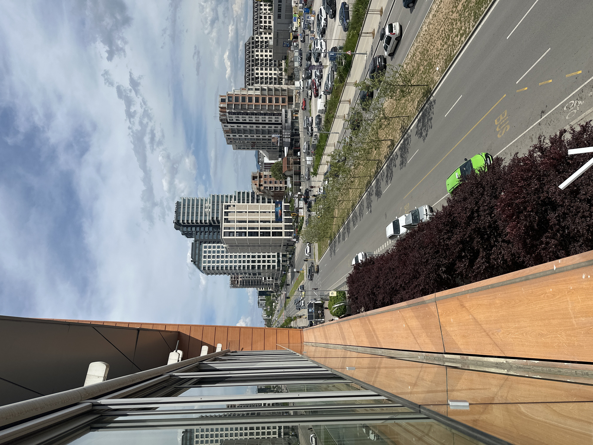 The image shows an elevated view of a street in Sofia, Bulgaria. High-rise buildings line the left side of the road. Traffic, including cars and a green bus, moves along the street. Trees are planted along the median and the sidewalk. A building is in the foreground, with a glass facade and wooden panels. The sky is visible with scattered clouds.