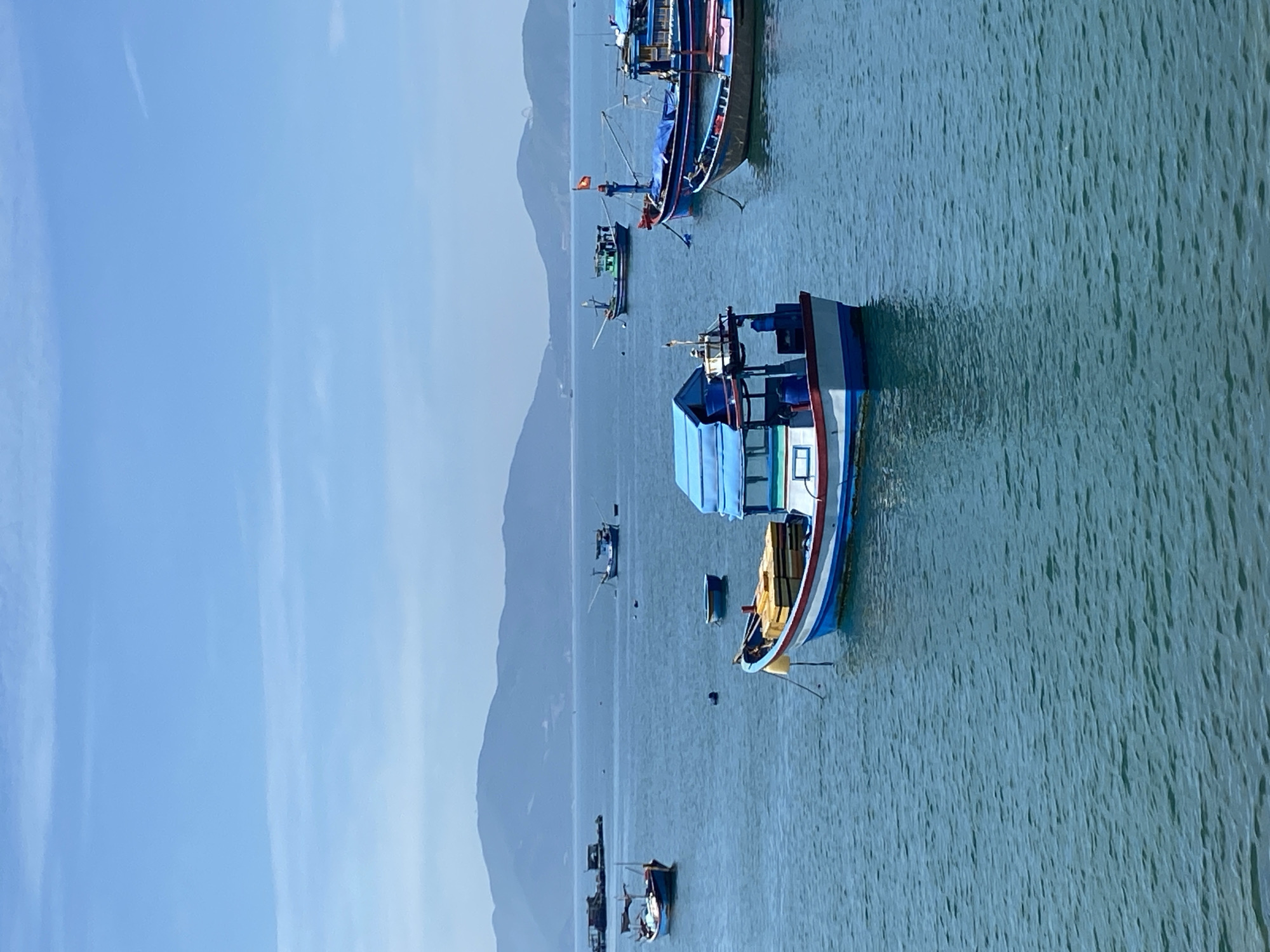 The image depicts a serene harbor in Nha Trang, Vietnam, on a clear, sunny day. The azure water is calm, with gentle ripples reflecting the bright sky. Several fishing boats of varying sizes are anchored in the bay. In the foreground, a colorful boat with a blue canopy and white and red trim is prominently featured. It carries a stack of yellow crates. Further out, other boats are visible, some with their masts and rigging, suggesting they are either docked or preparing for a fishing expedition. In the background, a range of lush green mountains form a picturesque backdrop against the vast expanse of the sky. The overall atmosphere is tranquil, showcasing a typical scene of coastal life in Nha Trang.