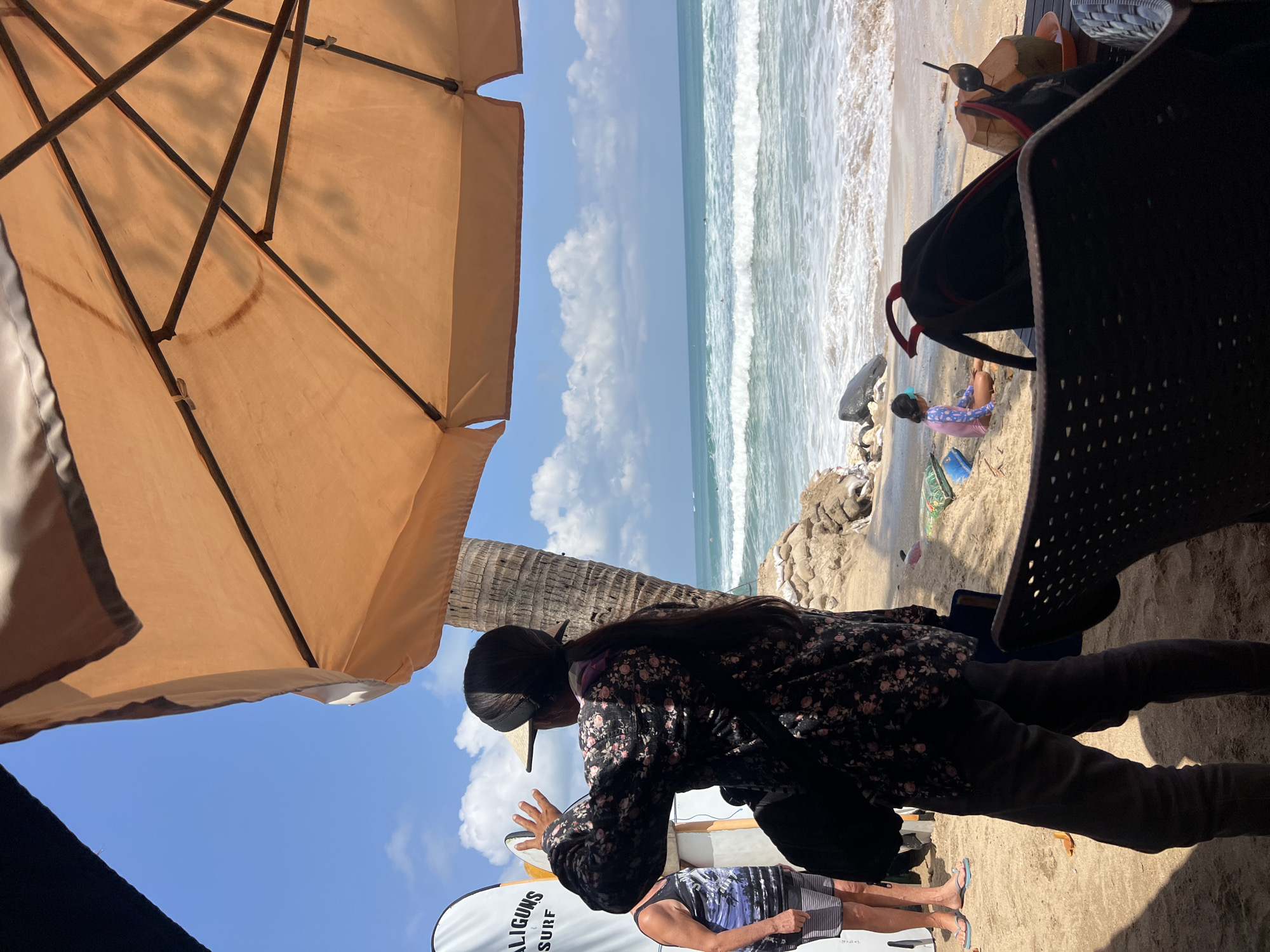 This image depicts a beach scene in Kuta, Indonesia, under a bright, sunny sky with scattered clouds. A large, orange beach umbrella provides shade in the foreground, covering part of the sandy shore. In the middle ground, a person wearing a dark, floral-patterned shirt and dark pants stands with their back to the camera, gesturing with their right hand as if interacting with someone or something out of frame. They are wearing a visor and have their hair pulled back. To the left, another person wearing a patterned tank top and striped shorts is visible.
Further down the beach, a child in a pink swimsuit sits on the sand, facing the ocean. The ocean itself is calm with gentle waves breaking on the shore. The beach is lined with sandbags and some rocks. In the immediate foreground, a dark, woven chair is positioned, and on a table to the right, a coconut with a straw and spoon is present. Surfboards are leaning against a structure on the left side of the image. The overall atmosphere suggests a relaxed, tropical vacation setting.