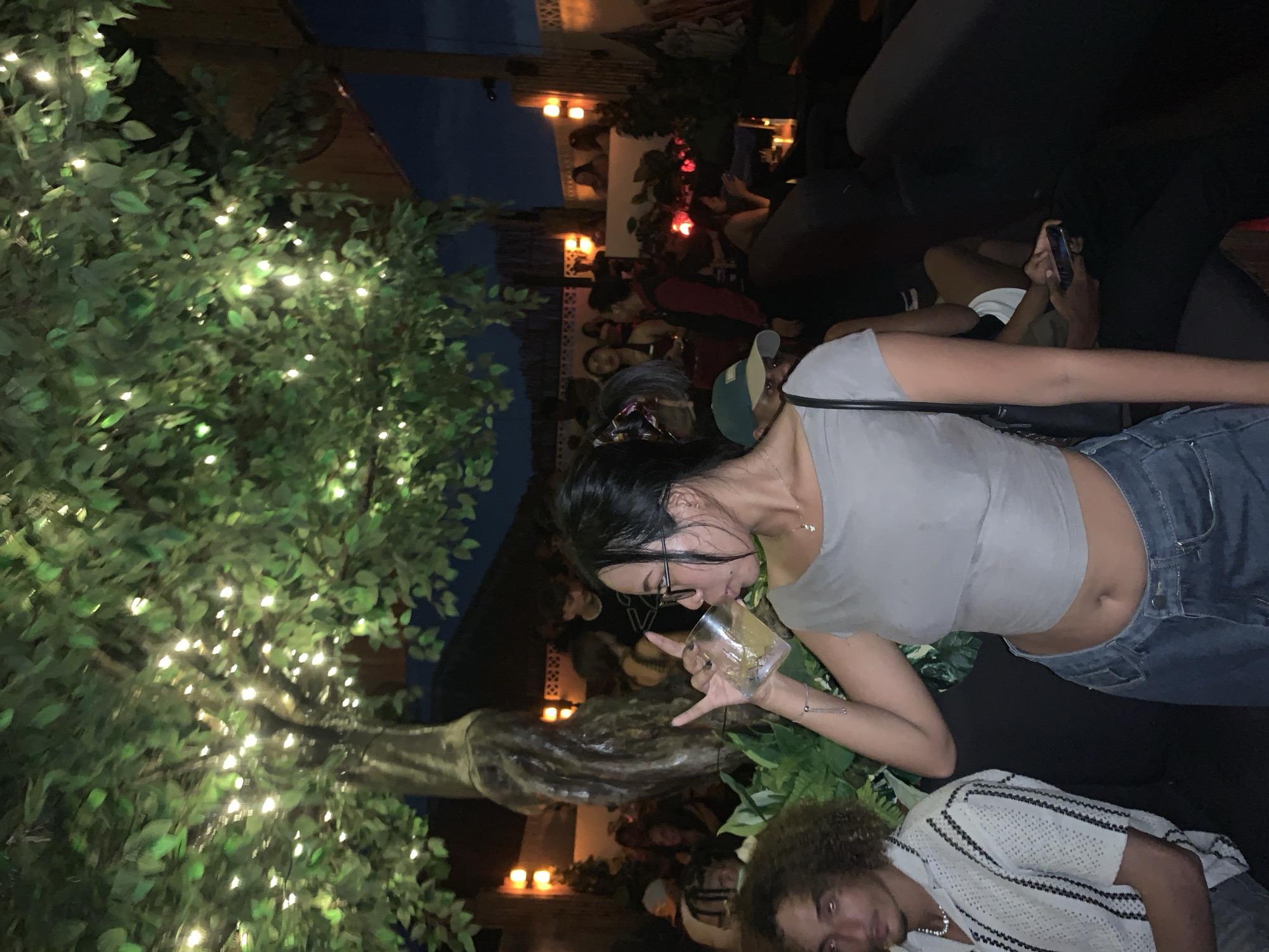 The image depicts an outdoor setting at night, likely a bar or lounge area, located in North Kuta, Indonesia. A woman with long, dark hair, wearing glasses, a grey crop top, and blue jeans, is drinking from a clear glass and making a hand gesture. A man with curly hair is visible near the bottom edge of the frame, wearing a white and black striped shirt. Several other individuals are seated in the background. The environment includes trees adorned with numerous small lights, indicating a festive or decorative atmosphere. Some are holding phones. Interior structure and architectural features are also seen.