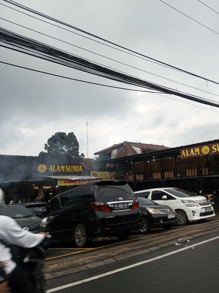 The image depicts a roadside scene in Dawathang_Dorjibi_ Kashingtsawa, Bhutan, under an overcast sky dominated by dense grey clouds. Numerous black utility cables are strung horizontally across the upper portion of the frame.
In the foreground, a paved road is visible with a white solid line marking its right edge and yellow parking bay lines further back. A person wearing a white helmet and white attire is partially visible on a motorcycle in the lower-left, moving from left to right.
To the right of the road, a line of vehicles is parked. These include a dark-colored SUV/MPV, a black Toyota Alphard with license plate B 1096 BB, a dark-colored sedan (likely a Nissan Livina), and a white Toyota Alphard with license plate D 1083 AAN (partially visible). Several other motorcycles are also parked further to the right.
Two prominent commercial buildings with the name "ALAM SUNDA" are central to the scene. The left building is a single-story structure with a dark roof and a large yellow "ALAM SUNDA" sign, from which smoke or steam is actively rising. Patrons are visible seated inside this establishment. The right building is a multi-story structure featuring a reddish-brown tiled roof and an open-air upper dining area. It also displays a large yellow "ALAM SUNDA" sign and a smaller sign stating "TASTE SENSATION Like No Other". A tall, slender antenna or tower is visible in the background, positioned between the two buildings, behind a large green tree.