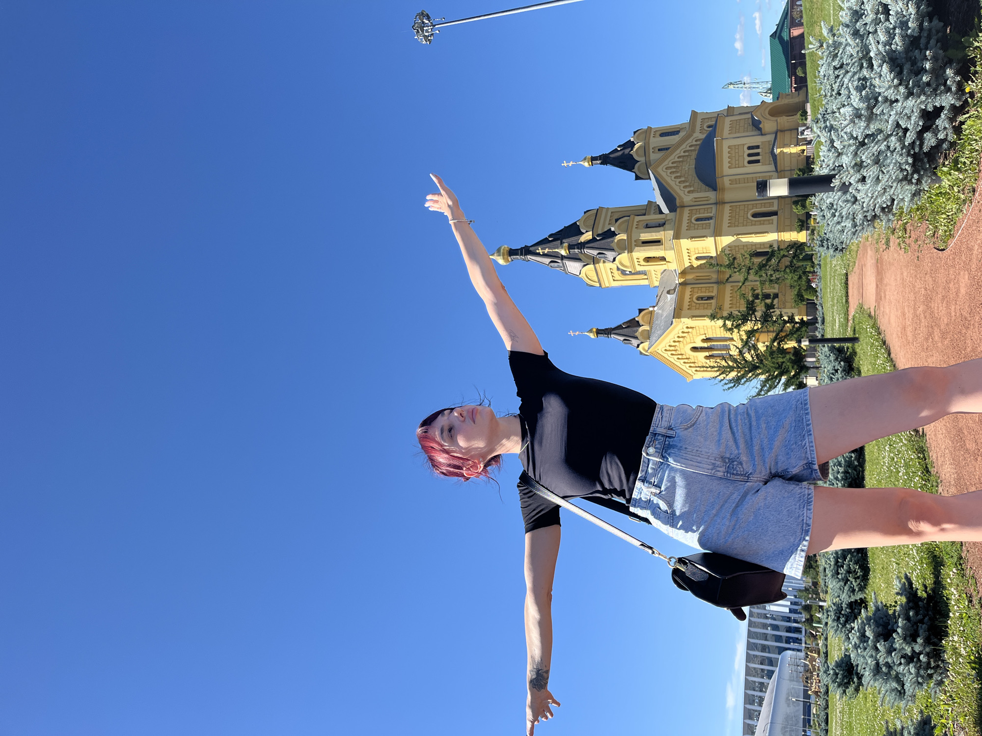 A young woman with dyed red hair stands with her arms outstretched against a clear blue sky in front of the Alexander Nevsky Cathedral in Nizhny Novgorod, Russia. She is wearing a black t-shirt and denim shorts, and a black crossbody bag is slung over her shoulder. The cathedral, a prominent landmark, is a bright yellow building with dark domes and ornate architectural details. The foreground features manicured green grass with scattered white flowers and neatly trimmed blue spruce bushes. A reddish-brown pathway winds through the grassy area, leading towards the cathedral. The scene appears to be set on a sunny day, with ample natural light illuminating the environment.