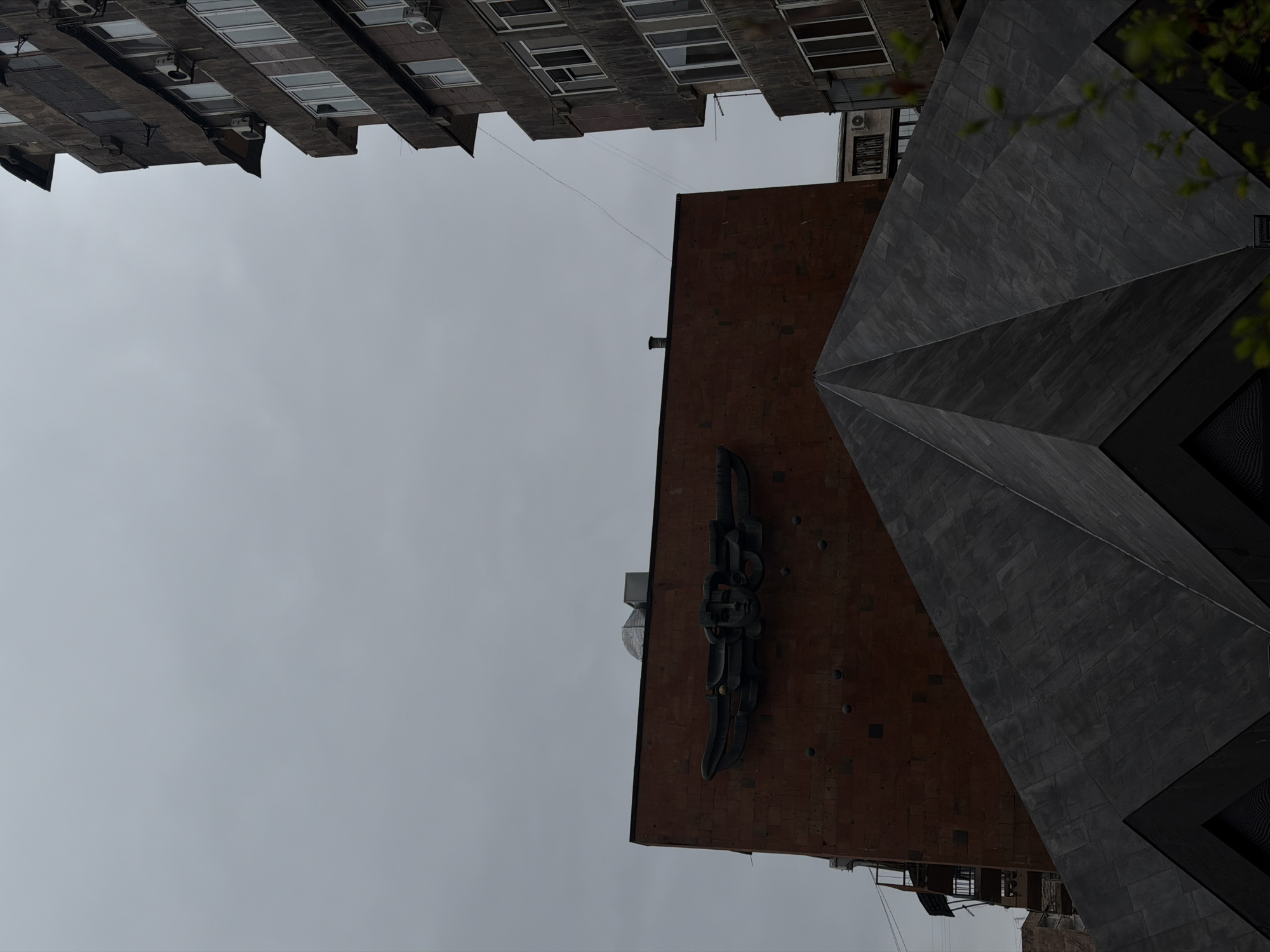 The image displays an urban scene in Yerevan, Armenia, featuring the Dvin Hotel at Paronyan St 40. An overcast, uniform light grey sky occupies the upper portion of the frame. In the central midground, a large, rectangular building facade of reddish-brown brick is prominent. Mounted on this facade, approximately one-third down from the top edge, is a dark, metallic, stylized sculpture identified as the "Winged Goddess of Victory," depicting a winged head. The brickwork exhibits subtle variations in color and texture, with some darker square and circular markings.
In the lower foreground and mid-center, a dark, angular, pyramid-like structure, likely a modern roof or architectural element, extends upwards and outwards from the bottom edge, obscuring the lower part of the brick building. This structure is composed of multiple triangular, dark grey facets.
To the right side of the image, a tall, narrow, multi-story building with a weathered, dark grey, possibly concrete or stone, facade extends from the mid-frame to the top. This building features multiple rectangular windows with white frames and numerous external air conditioning units attached below the windows and on small protrusions. Thin black cables or wires are visible traversing the open sky between the reddish-brown building and the multi-story building on the right. Green foliage is partially visible in the lower right corner of the frame. No individuals are present in the scene.