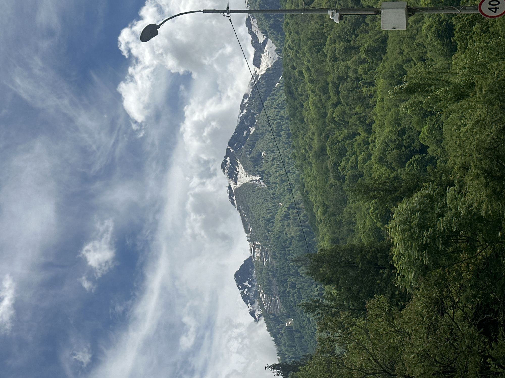The image shows a mountainous landscape in Krasnaya Polyana, Russia. The mountain is partially covered with snow, especially at higher altitudes. A dense green forest covers the lower slopes. The sky is blue with scattered white clouds. A street lamp is visible in the foreground, along with a sign indicating a speed limit of 40. Power lines run through the scene. The photograph appears to have been taken during the daytime.