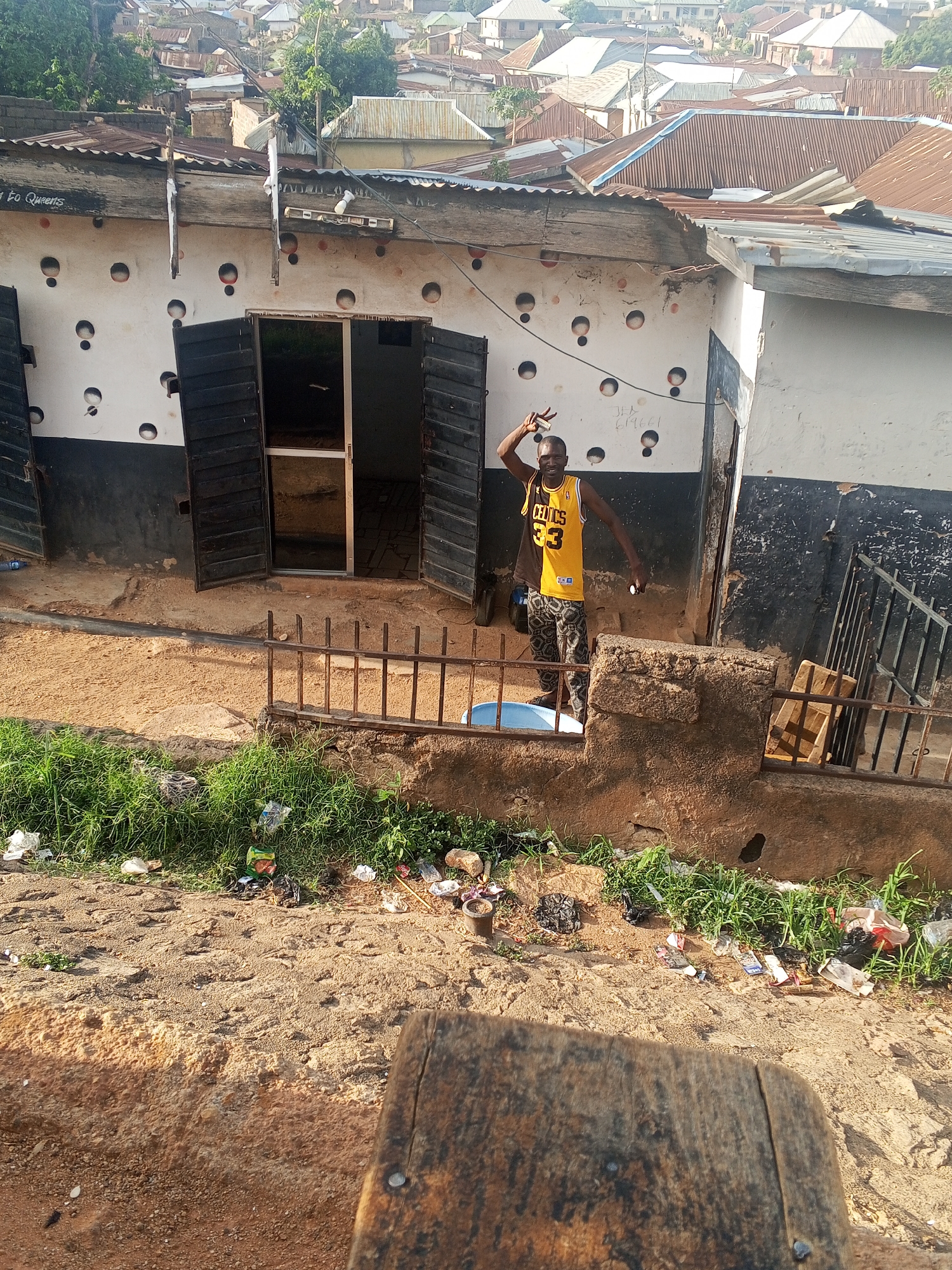 A man wearing a yellow and black Celtics #33 jersey stands in the doorway of a building with white walls decorated with black circles in Gwong, Nigeria. He holds up two fingers in a peace sign with his left hand and what appears to be a small white object in his right hand. The building has an open door and open black shutters on either side. A low, rusty metal fence runs in front of the building. Below the fence, there is a patch of green vegetation with scattered litter. In the foreground, there is a blurry wooden object, partially obscuring the view. In the background, there are densely packed buildings with corrugated metal roofs.