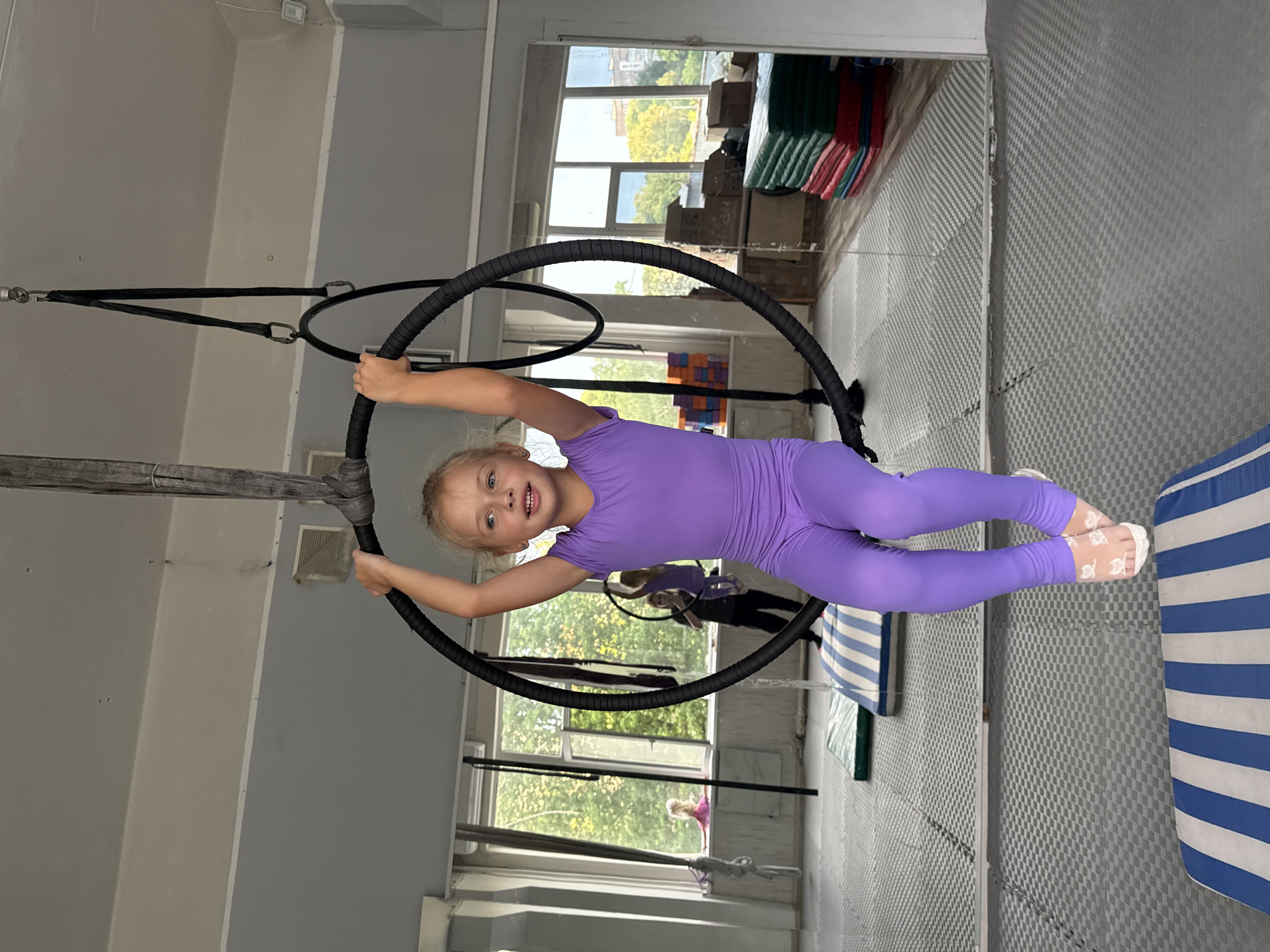 A young girl with blonde hair, wearing a purple leotard and white socks with a butterfly pattern, is suspended mid-air within a black hoop. She smiles widely at the camera. The hoop is attached to the ceiling by black ropes and carabiners. The scene appears to be an indoor gymnasium or studio, suggested by the grey walls, checkered floor mats, and large windows showing greenery and some stacked boxes and mats in the background. There are also other hoops visible, as well as other people in the background, one of whom also appears to be engaged in aerial activities. The lighting suggests it is daytime. The overall mood is energetic and joyful. The location is Dnipro, Ukraine.