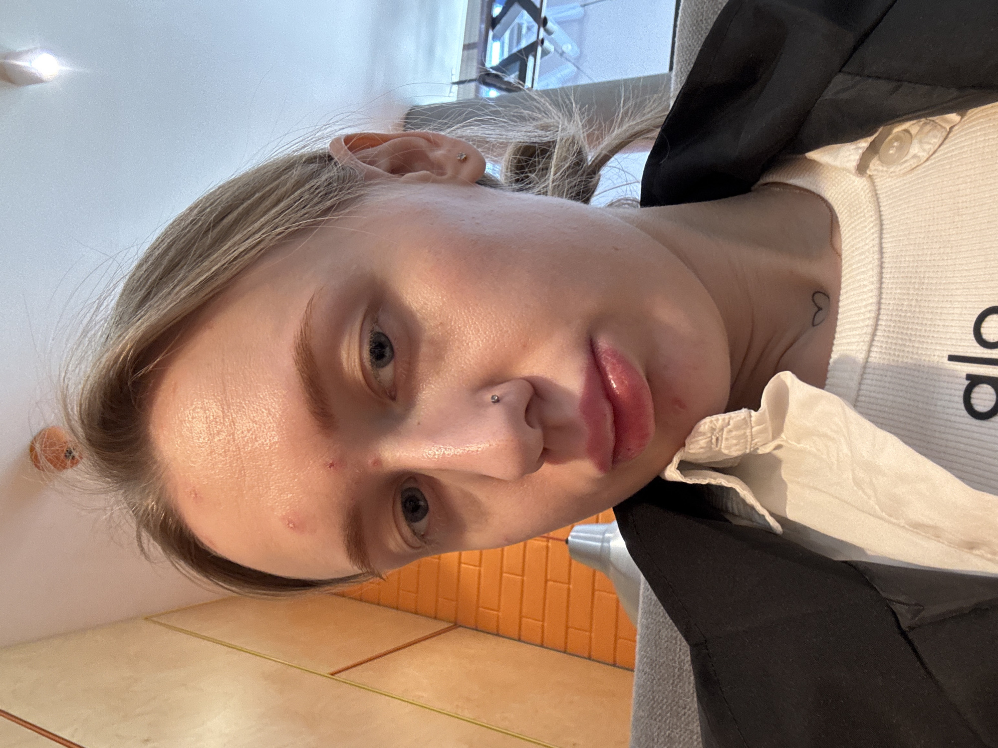 A close-up selfie shows a woman with fair skin, blonde hair, and blue eyes. A small stud is visible in her left nostril. She has a few minor blemishes on her forehead. Her lips are full and slightly parted. She wears a white knitted sweater with "alo" printed on the front, layered under a dark-colored garment. A small heart tattoo is visible on her neck, near the collarbone. The background includes orange and yellow tiled walls. The location is not discernible to be Rostov-on-Don based on the image alone.