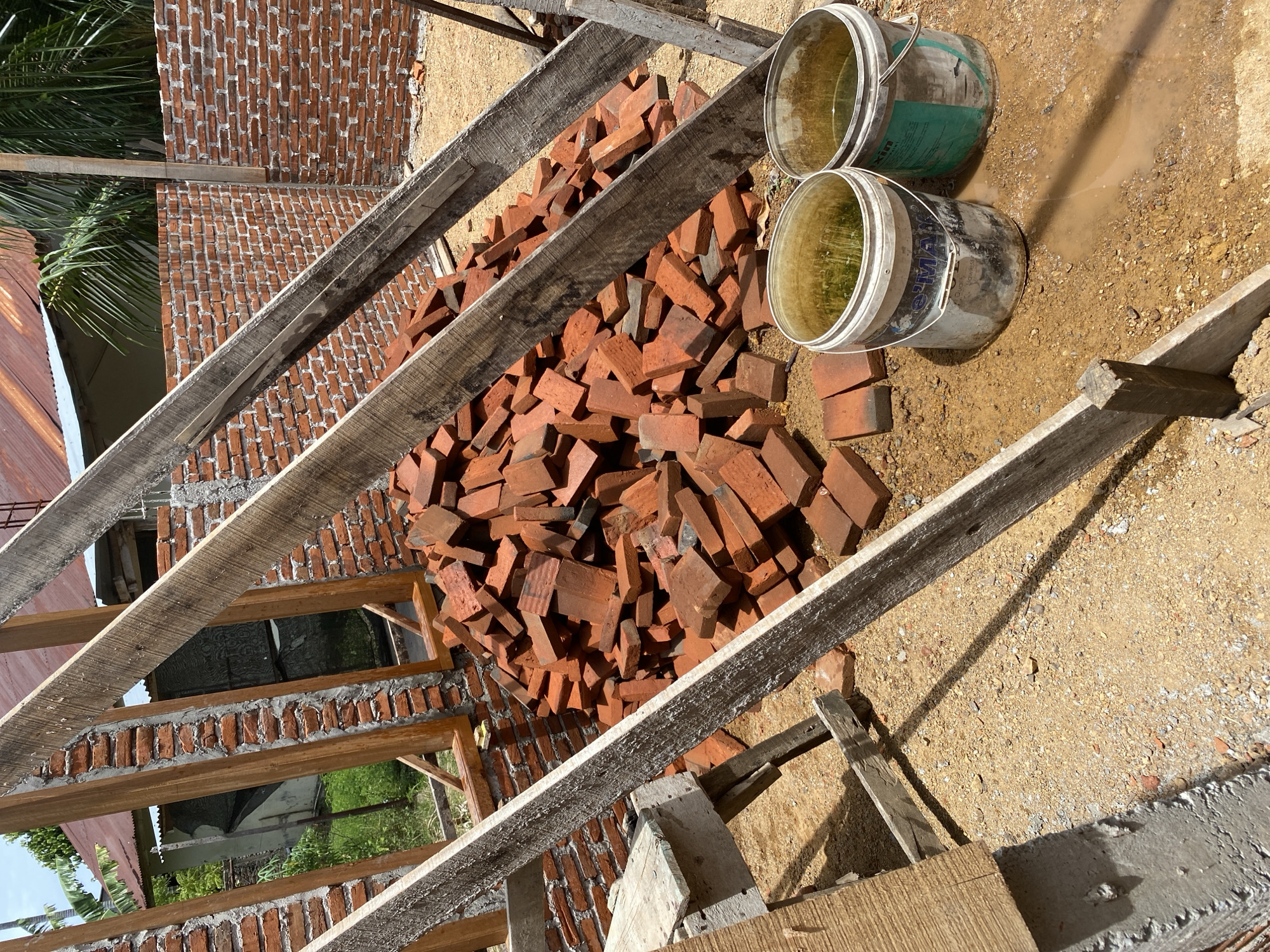 A construction site is depicted with a large pile of red bricks in the center of the frame. Wooden beams, likely part of a scaffolding or support structure, crisscross the scene, casting shadows on the dusty ground. Two buckets, one white and one gray, are placed on the ground near the bricks, both containing a murky liquid. In the background, unfinished brick walls are visible, along with a partial view of a building with a corrugated metal roof and some greenery. The overall impression is one of ongoing construction activity in an outdoor setting.