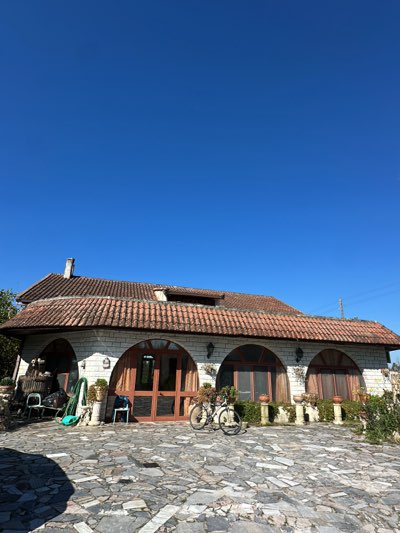 The image depicts a building with a beige facade, a red-tiled roof, and arched doorways, standing on a cobbled surface in Cutrofiano, Italy. A bicycle rests in front of the central doorway. The sky is clear and blue. The scene is captured in daylight. The architecture appears to be residential.