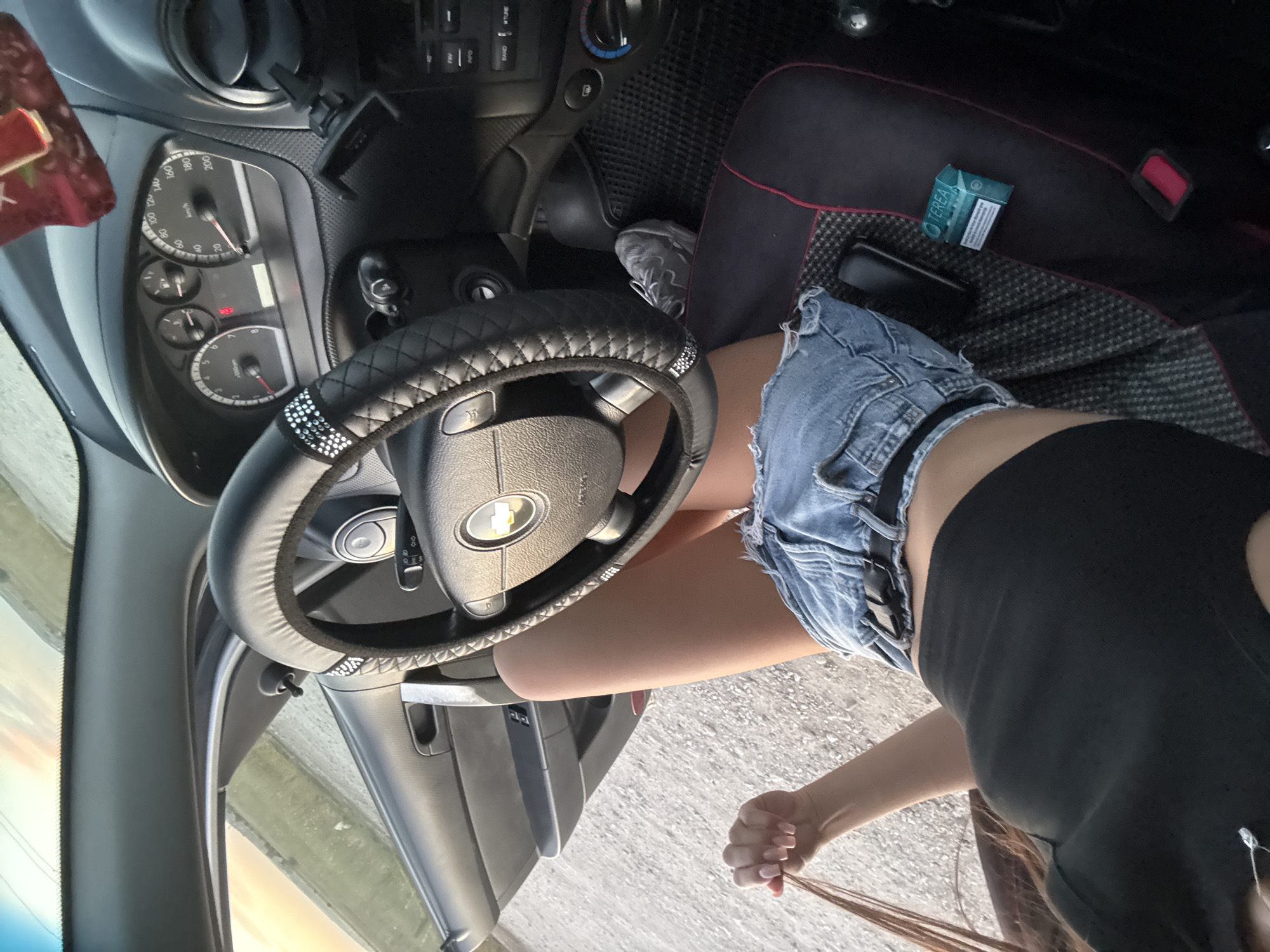 The image shows a close-up view inside a car, focusing on the driver's seat area. A person, partially visible, is sitting in the driver's seat. They are wearing a black crop top and distressed denim shorts. The person's right hand is pulling on their hair. A steering wheel, covered in a black material with diamond-like accents, is prominently displayed. The dashboard instruments are visible, including a speedometer. A pack of TEREA cigarettes is on the seat. The car appears to be parked on a gravel surface, with a rural landscape visible through the open car door and windshield. The lighting suggests it is daytime, possibly late afternoon given the warm tones of the sky. The location is identified as Cimișlia, Moldova.