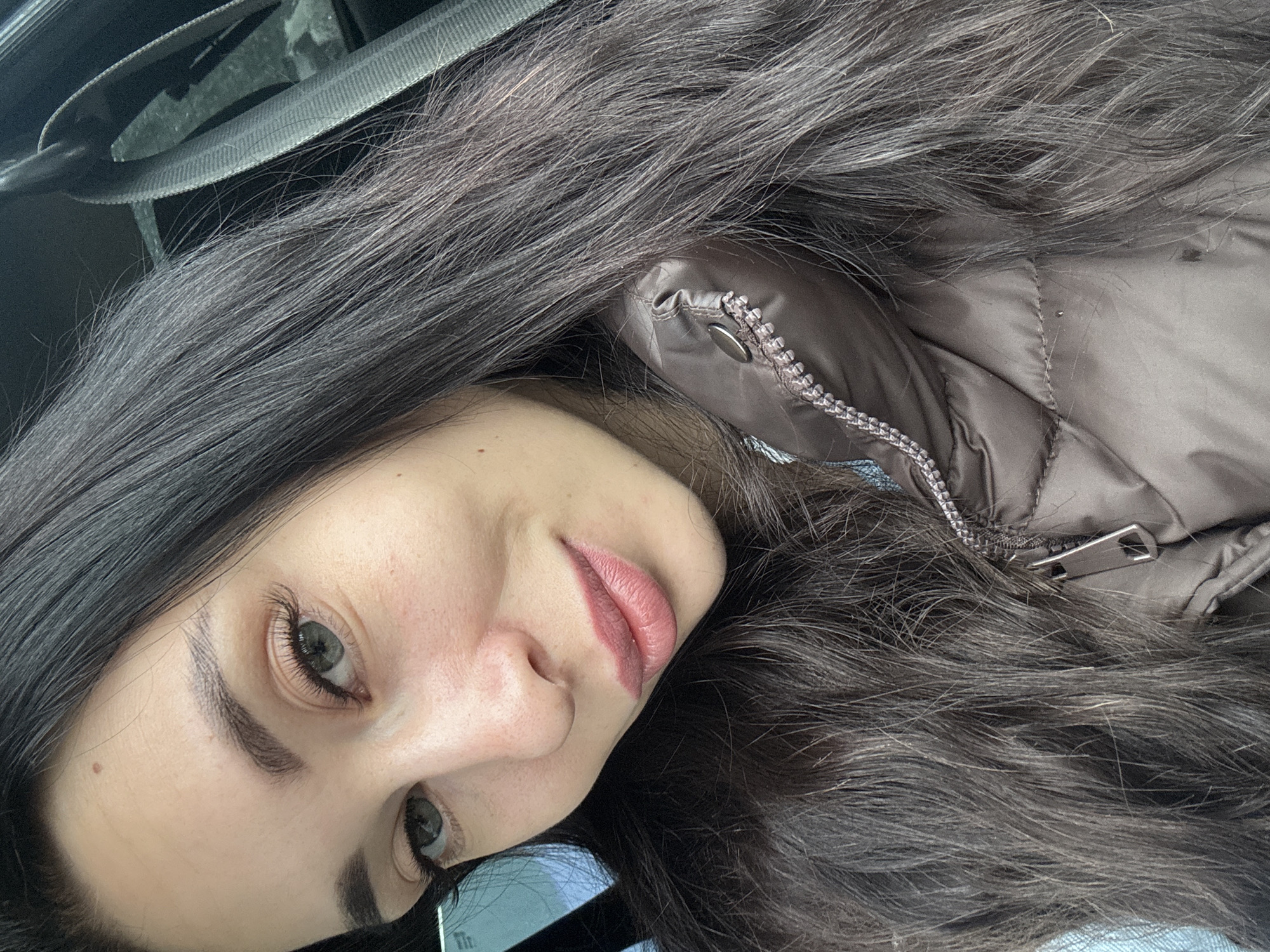 A selfie of a woman with long dark hair, light skin, and neutral-toned makeup. She is wearing a brown puffer jacket. The image appears to be taken inside a vehicle, as a seatbelt strap is visible in the upper left corner. No information to suggest a location in the city of Chaikovsky, Russia.