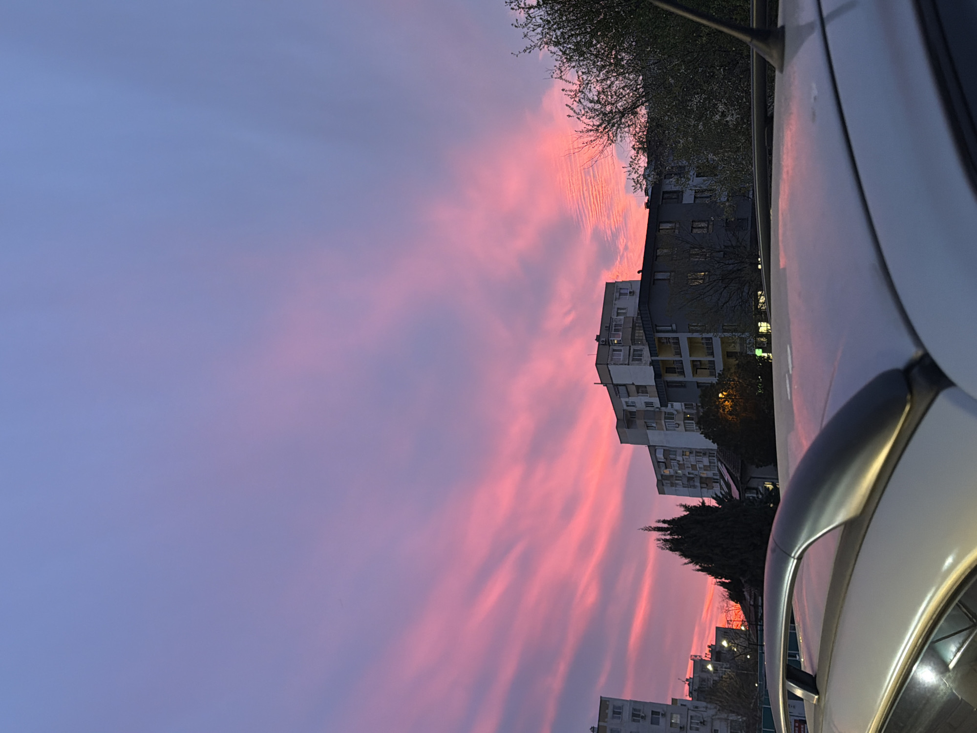 The image captures a vibrant sunset in Varna, Bulgaria. The sky transitions from blue to pink, with striated clouds illuminated by the setting sun. The foreground includes a partial view of a white car, likely parked on a street. In the background are several apartment buildings, typical of urban residential areas in Varna. Trees are interspersed between the buildings. The overall scene suggests a quiet evening in a residential neighborhood.