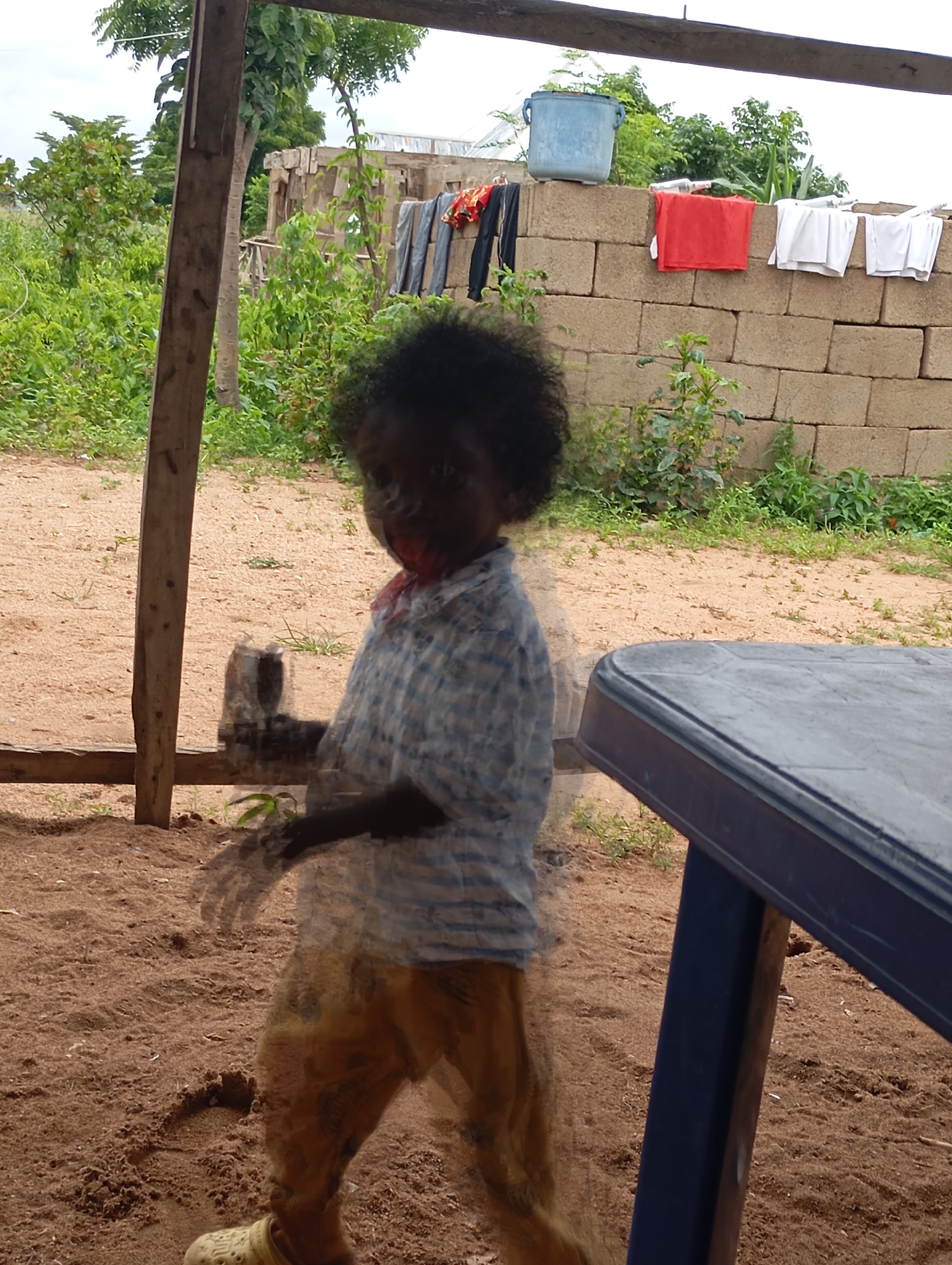 The image depicts an outdoor scene, likely in a rural or semi-urban area. The ground is covered in light brown sand and scattered organic debris. A child, appearing to be of African descent with dark, curly hair, is the central subject. The child is wearing a light blue and white checkered shirt and light brown pants. The child's movement is blurred, suggesting motion within the frame.
In the background, a wall constructed from beige concrete blocks is visible. On top of this wall, several items of clothing are hung out to dry: a red cloth, and two stacks of white cloth. To the left of the clothing, a large, light blue plastic bucket is placed. Beyond the wall, lush green vegetation and trees are abundant, indicating a natural environment. The sky appears overcast.
To the right of the child, a dark blue plastic table is partially visible. In the foreground, a wooden support structure, likely part of a shelter or building, frames the left side of the image. The overall impression is one of a domestic or communal outdoor space. The image was taken in Shendam, Nigeria.