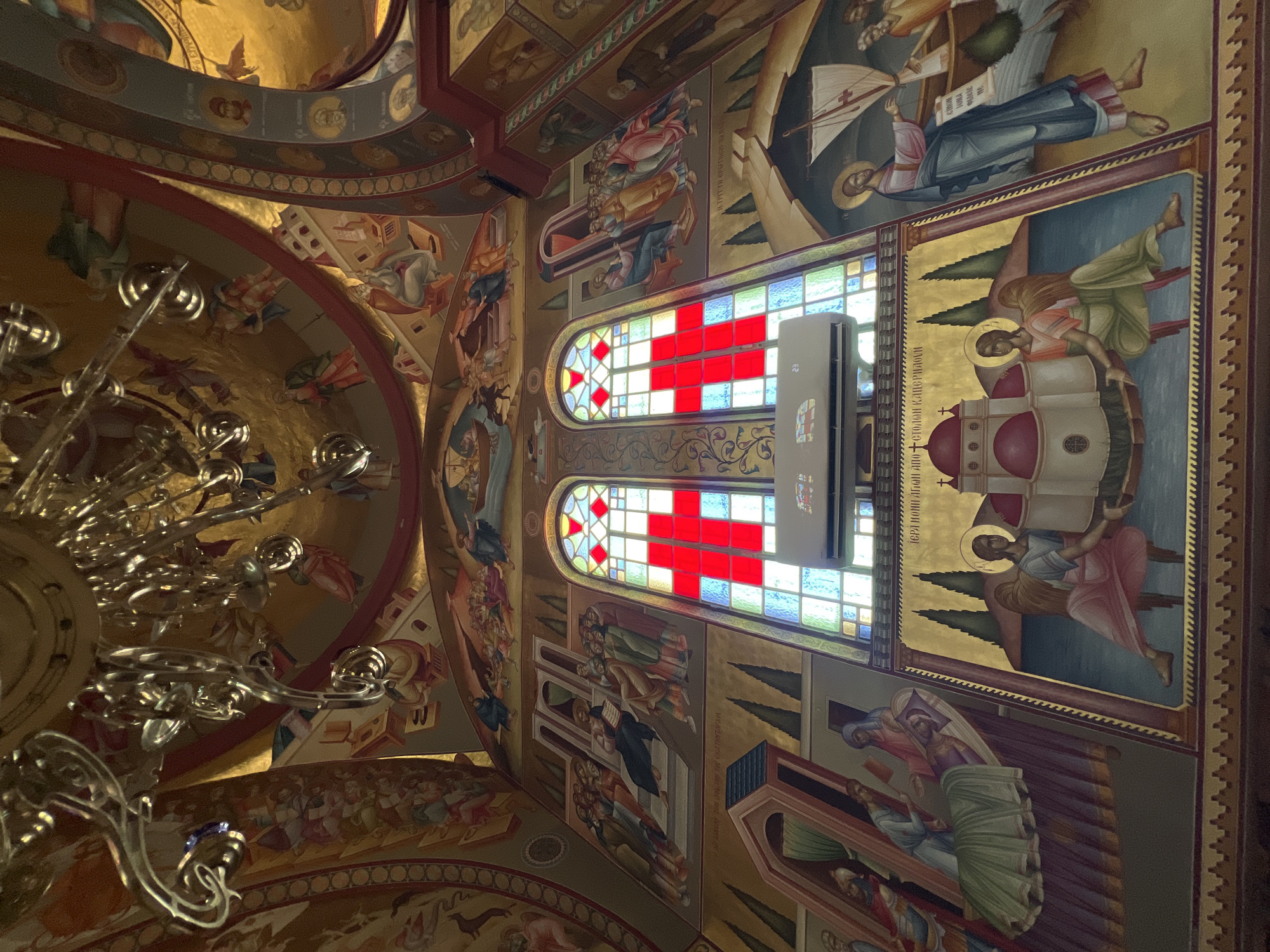 The image depicts the interior of a church, likely in Emek HaYarden Regional Council, Israel, with ornate religious frescoes adorning the walls and ceiling. A large, multi-tiered chandelier hangs from the ceiling, reflecting light. The walls are covered with detailed paintings of biblical scenes and figures, rendered in vibrant colors with gold accents. In the center, two stained-glass windows are prominently featured, each illuminated by red and white patterns that resemble crosses. Below the windows, an air conditioning unit is installed. The lower portion of the walls displays further religious imagery, including depictions of angels, religious structures, and figures interacting with each other. The overall impression is one of rich artistic decoration and spiritual significance within the church setting.