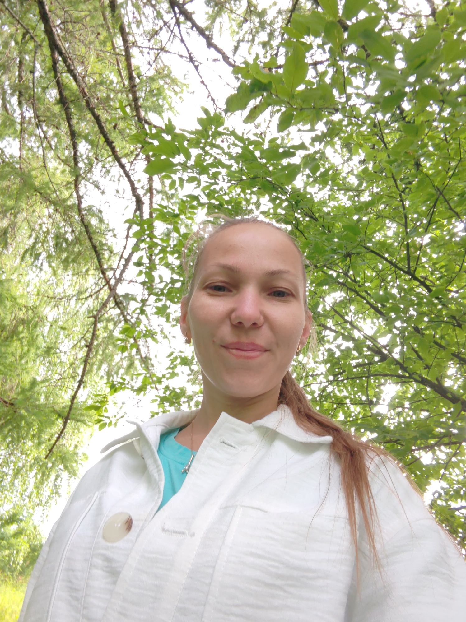A woman with light brown hair pulled back is photographed from a low angle, looking up at the camera. She is wearing a white jacket over a turquoise shirt. Her face is centered in the frame, and she has a slight smile. The background is dominated by the bright green leaves and branches of trees, suggesting an outdoor setting. The sky is visible through the foliage. The image appears to be taken on a clear day. The location is specified as Chukhlem, Russia.