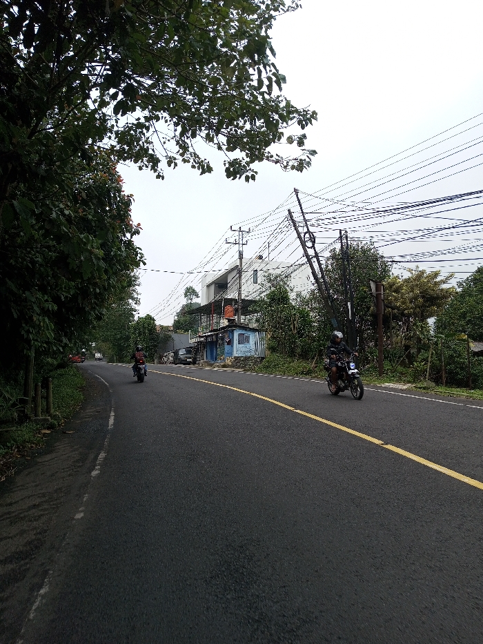 An outdoor scene in Neerveli, Sri Lanka, depicts a paved asphalt road under an overcast sky. The road features a solid yellow center line and a faded white edge line on the left, curving slightly into the distance. Lush green foliage and trees line both sides of the road, with a large tree canopy extending into the upper left quadrant.
In the foreground to mid-ground, two individuals are operating motorcycles. One motorcyclist, wearing a dark helmet and clothing, rides a dark motorcycle with a white headlight housing towards the viewer on the right side of the road. A second motorcyclist, similarly attired, rides a dark motorcycle away from the viewer on the left side of the road.
On the right side of the road, a cluster of utility poles supporting numerous overhead wires is visible. One pole appears braced or leaning diagonally. Behind the utility poles and dense vegetation, a modern, multi-story white building with visible windows stands on an elevated position. Below this building and closer to the road, a light blue, corrugated shed-like structure is present, flanked by a darker, open-air commercial or stall-like structure. Several dark-colored vehicles are parked near these structures. A faint red vehicle is visible in the distant background on the left side of the road. The road surface appears dark and possibly damp.