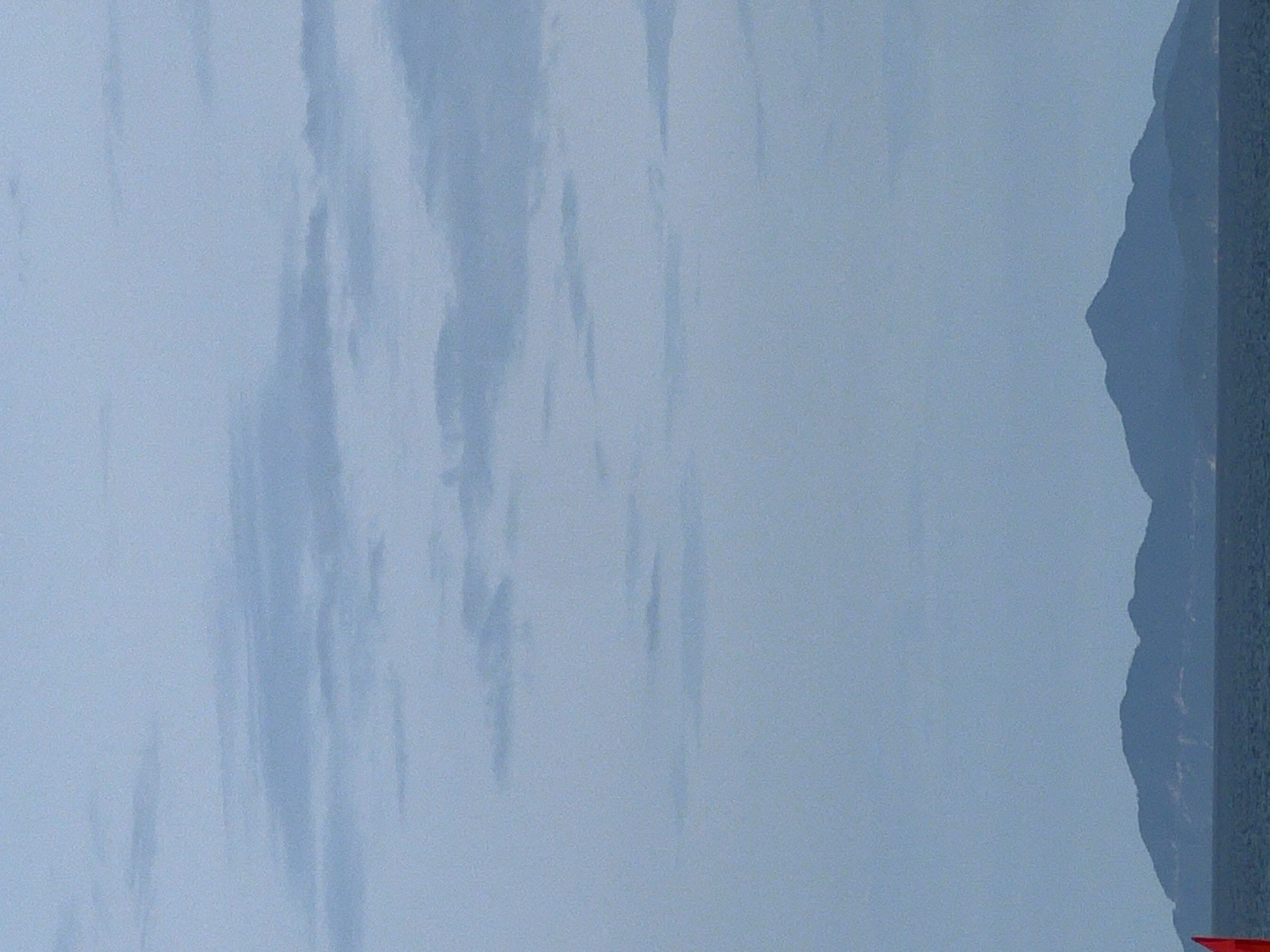 The image depicts a serene seascape with a distant mountainous island range under a hazy, overcast sky. The foreground is dominated by a calm, dark blue sea, and to the far left, a portion of a bright red flag is visible, partially cropped out. The island range, appearing as dark, silhouetted shapes against the horizon, is characteristic of islands visible from the coast of Đà Nẵng, Vietnam, such as those in the Hoàng Sa (Paracel) Islands chain. The sky is a soft, pale blue with diffused, horizontal cloud formations, suggesting a potentially humid or overcast day. The overall atmosphere is tranquil and somewhat muted due to the atmospheric conditions. There are no people or discernible structures on the visible portion of the sea or the island.