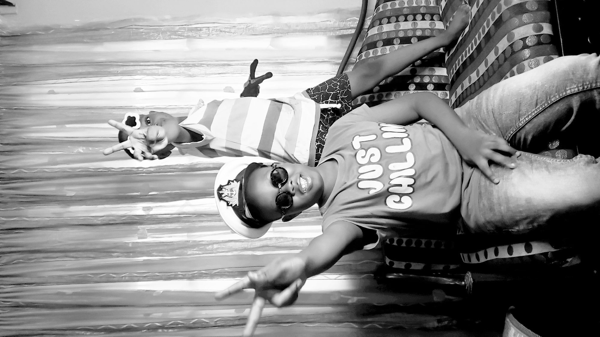 Two children lie on a bed. One wears a "Just Chillin" t-shirt, sunglasses, and a naval cap. The other wears a striped shirt. Both are making peace signs with their hands. The bed has patterned pillows and a striped bedspread. The location appears to be Narayi, Nigeria.