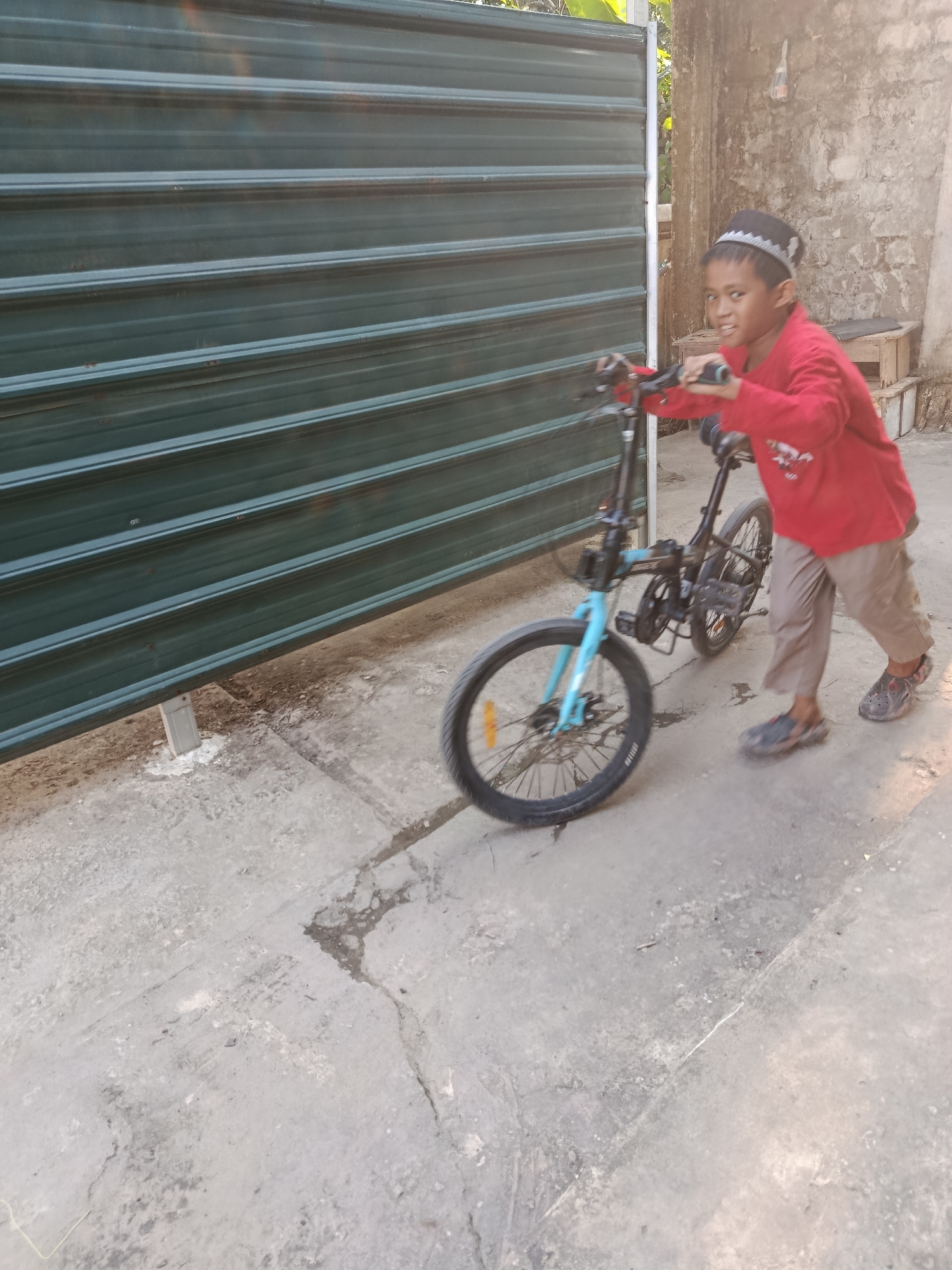 In Swarga Bara, Indonesia, a young boy wearing a red long-sleeved shirt and a black and white patterned cap pushes a folding bicycle. The boy is standing on a concrete surface with his left foot slightly forward. The bicycle is black and blue with visible spokes on the wheels. Behind the boy is a dark green corrugated metal wall that runs horizontally across the image. To the right of the boy, a rough, textured concrete wall is visible, with some wooden crates stacked against it. The scene appears to be outdoors, possibly in a residential or alleyway setting, with sunlight casting shadows.