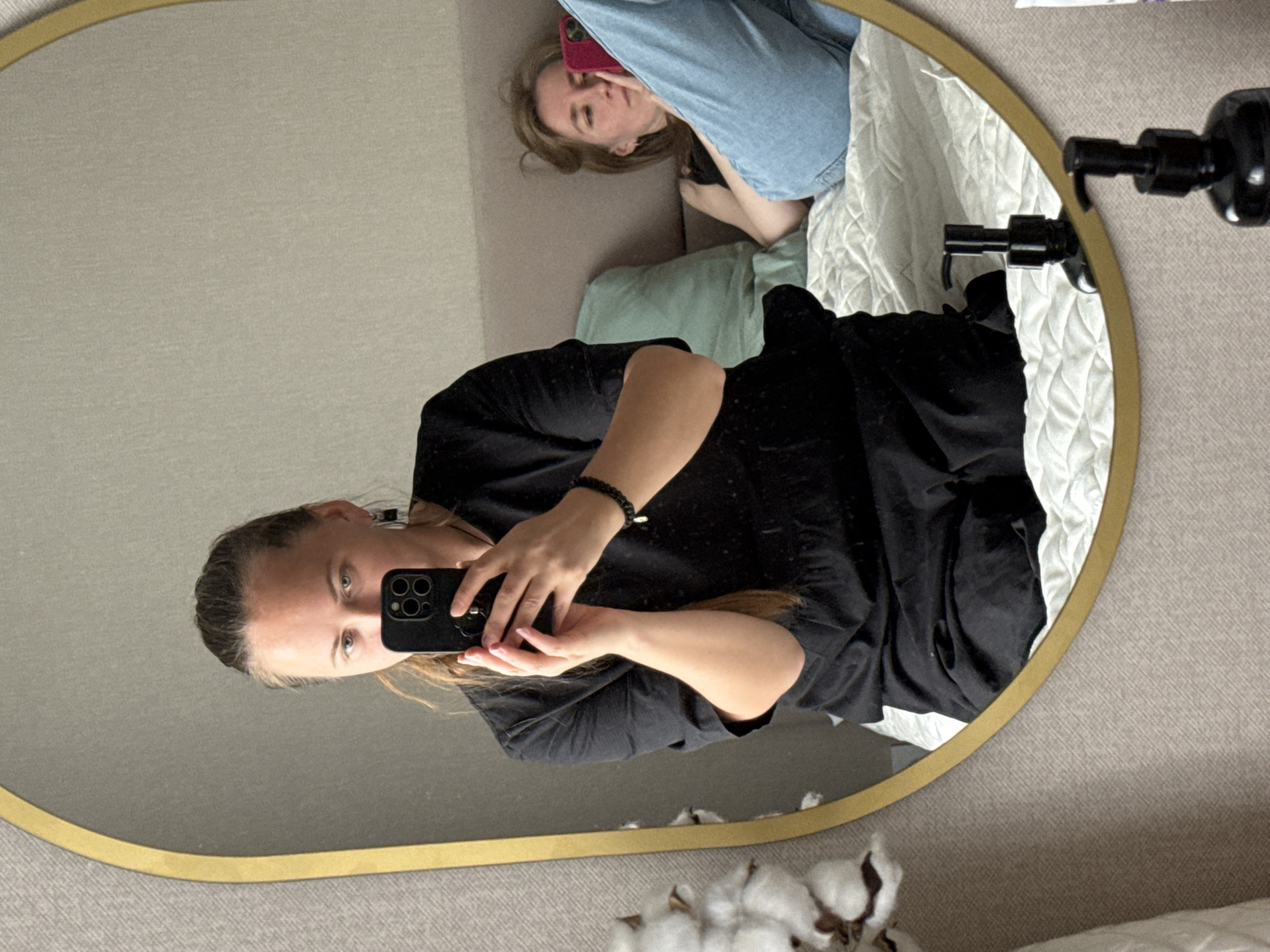 A young woman holds up a black smartphone to take a mirror selfie. The mirror's frame is gold and oval-shaped. The woman is wearing a black garment and a bracelet with a gold charm. Her hair is blonde. Reflected in the mirror, another woman with blonde hair is lying on a sofa and looking at a phone with a pink case. A portion of a black dispenser is visible near the top right corner of the mirror. The background shows beige walls and bedding. A few cotton plants are visible at the bottom. The scene is located indoors. There is no information to suggest this is located in the city of Izhevsk, Russia.