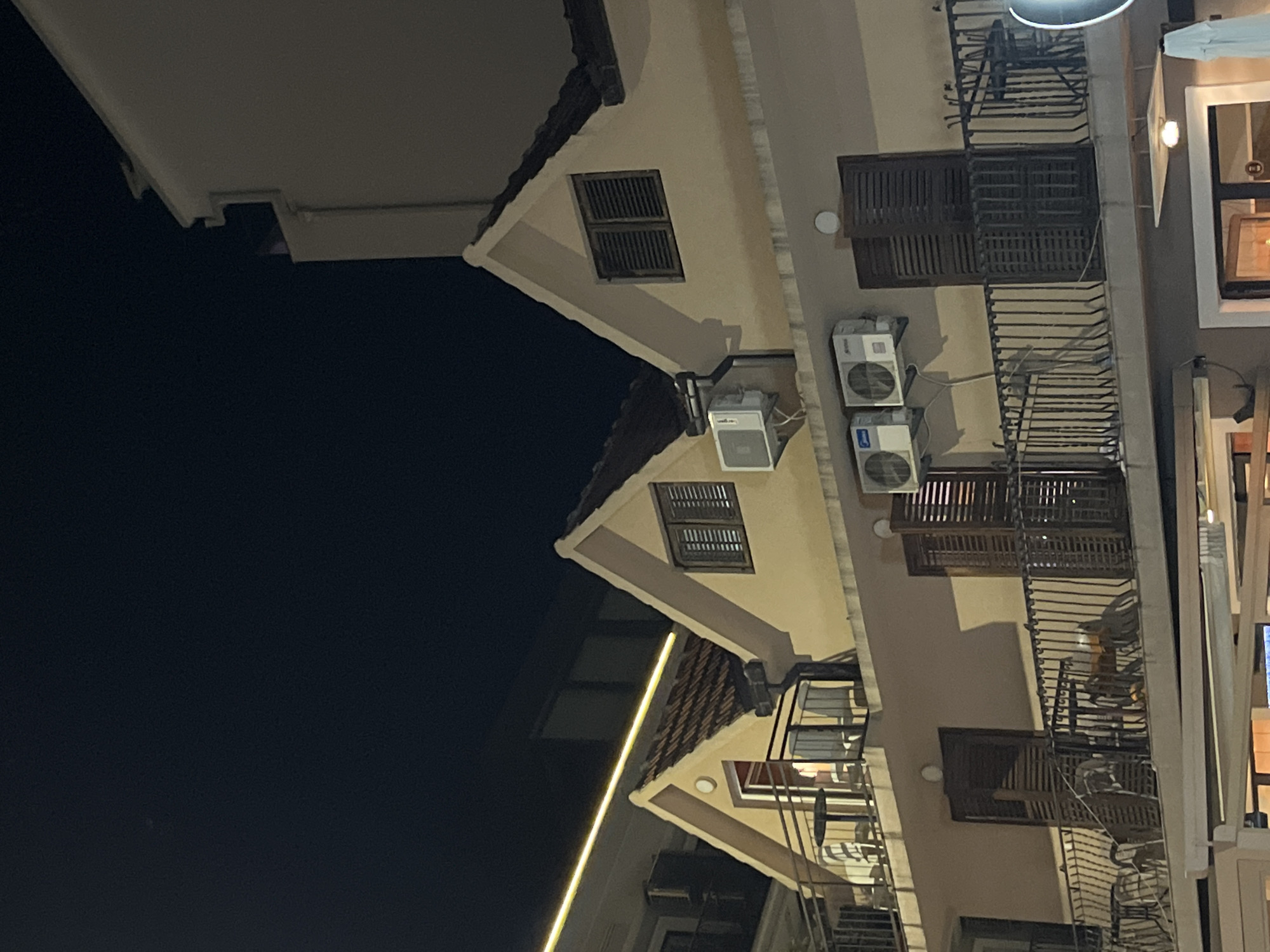 The image shows the exterior of a building in Budva, Montenegro at night. The building has a light beige facade with several pitched roofs topped with red tiles. There are multiple balconies with black metal railings. On the balconies, there are tables and chairs. Several windows with dark shutters are visible. Two air conditioning units are mounted on the building's facade. A bright yellow light strip runs horizontally across the upper part of the image, illuminating a section of the building and the dark night sky. The overall scene depicts residential or commercial units in a European coastal town at night.