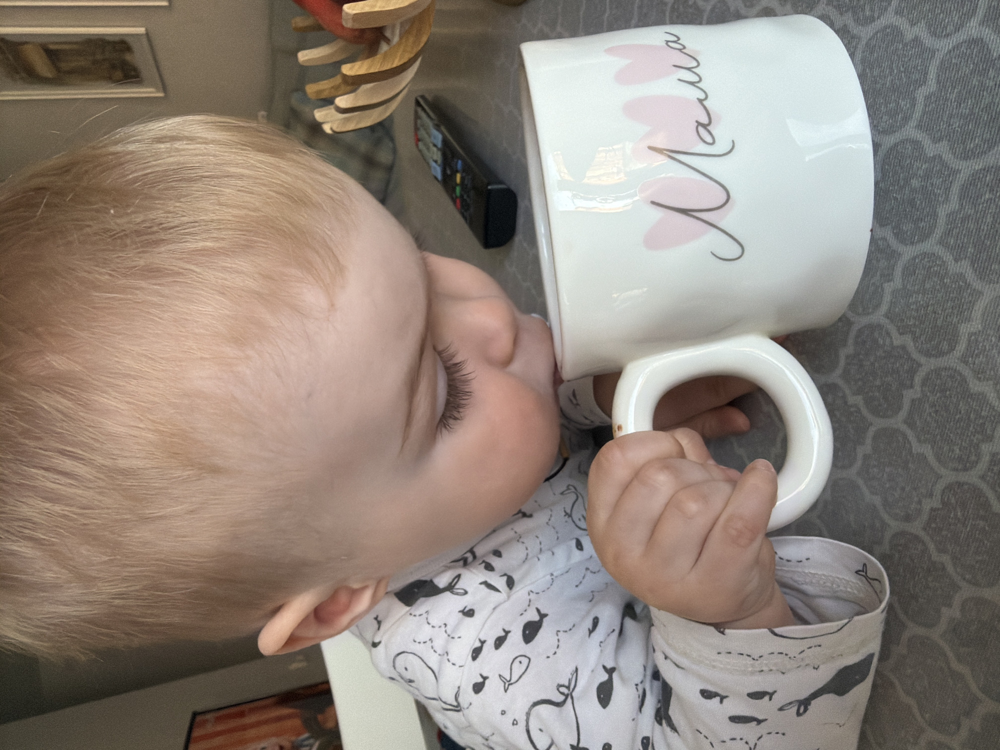 A baby with blond hair is drinking from a white mug. The mug has pink heart designs and the word "Mama" written on it in cursive. The baby is wearing a white pajama top with black whale designs. A black TV remote is visible next to a wooden object. The surface beneath is grey with a patterned design. The location is likely inside a home. There is no information in the image to determine a location in Votkinsk, Russia.