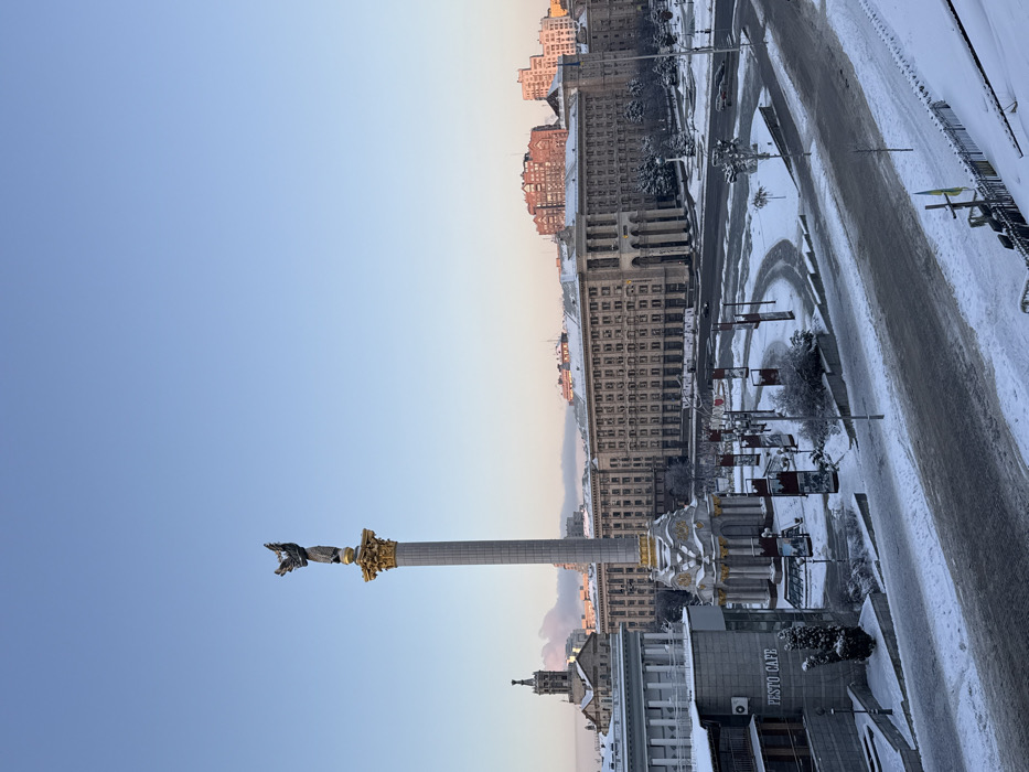 A city view during winter, likely Kyiv, Ukraine, features Independence Square. The iconic Independence Monument stands tall against a blue sky. Snow covers the surrounding buildings, streets, and sidewalks. Buildings line the square, some with visible signs like "Pesto Cafe." The cityscape is bathed in the soft light of either dawn or dusk. A street runs along the right side of the photo.