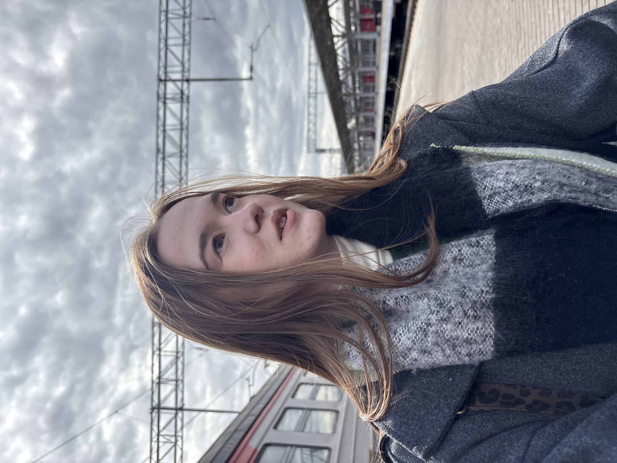 A woman is at a train station in Kazan, Russia. She wears a gray coat and scarf. An electric pylon and cloudy sky are visible above. A train is behind her.