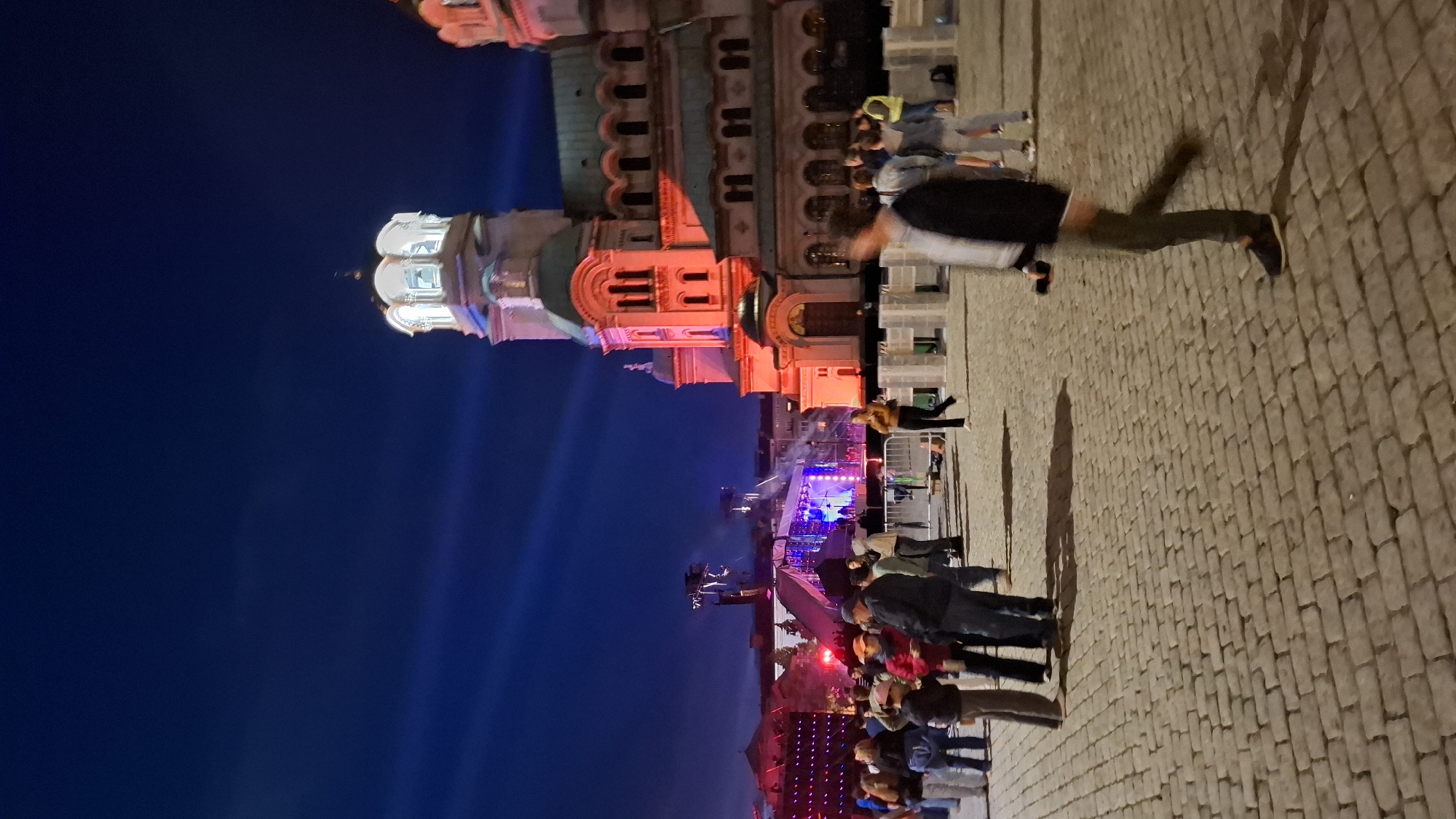 The image shows the Alexander Nevsky Cathedral in Sofia, Bulgaria, at night. The cathedral is illuminated with red and white lights. In the foreground, people are gathered on a cobblestone plaza, with a stage and lights visible in the background, suggesting an event is taking place. One person in the foreground is walking towards the right, with their face blurred. The sky is dark blue with blue lights projected upwards from the stage area.