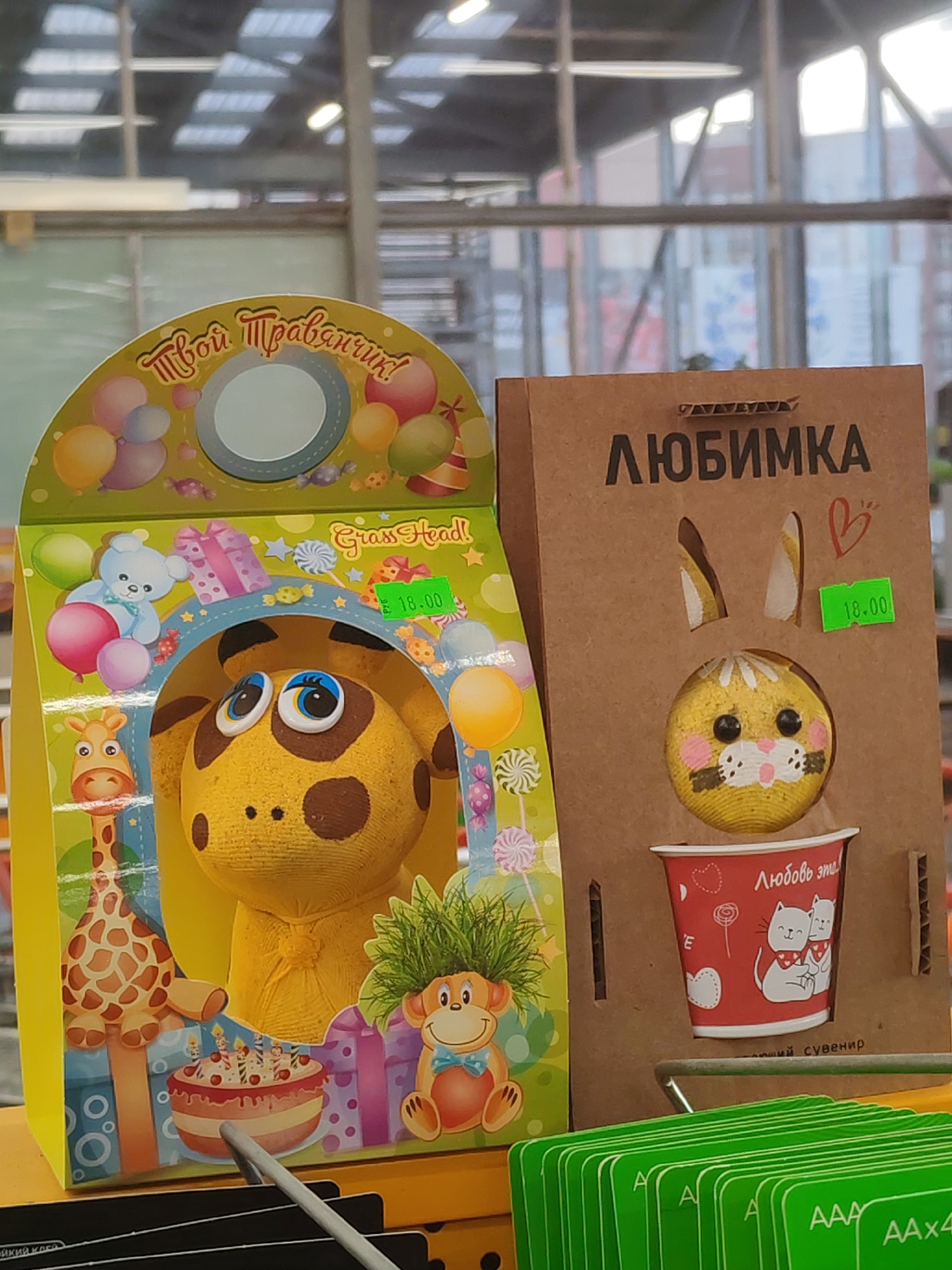 The image shows two packages of toys displayed for sale, likely within a retail setting. The package on the left features a cartoon giraffe and monkey, labeled "Grass Head!" with the price 18.00. The package on the right is a brown cardboard design displaying a cartoon bunny face above a cup, labeled "Любимка" (Lyubimka). The surrounding environment suggests an indoor location with blurred background elements. The green shelf is labeled AAA AAx4.