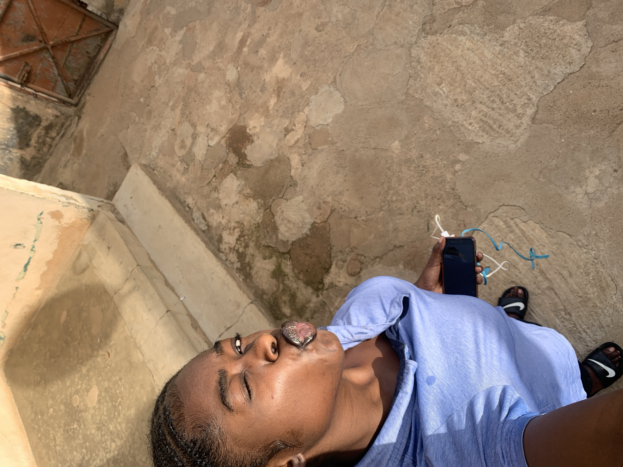 The image depicts a person lying down, seemingly on a concrete or stone surface. The person is dark-skinned, with braided hair. They are wearing a light blue button-down shirt. The person is holding a black smartphone in their right hand, with white earphones plugged in. They are also wearing black Nike sandals. The background is a textured concrete surface with some variations in color and possibly some cracks. In the upper left corner, there's a dark, rectangular object that could be a metal grate or a hatch. The location appears to be an outdoor area in Rafin Par, Nigeria.