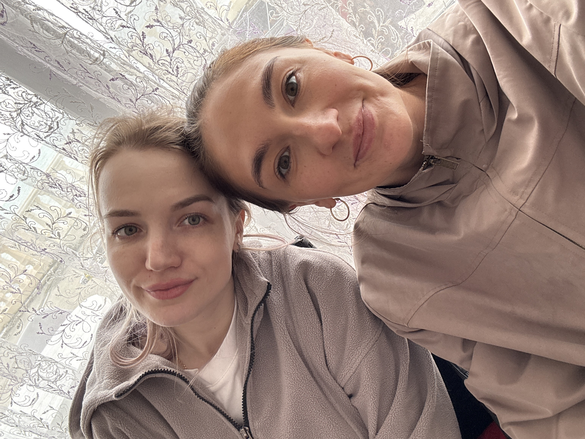Two women are captured in a close-up selfie, indoors in Sochi, Russia. The image is taken from a slightly upward angle, giving a casual, intimate feel.
On the left, a woman with fair skin, light-colored hair, and light eyes is seen from her forehead down to her torso. She wears a light grey or beige fleece jacket with a zipper, over a white t-shirt. Her expression is gentle, with a soft smile.
Leaning into her, on the right side of the frame, is another woman. She has darker hair pulled back, darker eyes, and is wearing a light brown or beige jacket with a zipper, possibly made of a smooth, slightly shiny fabric. She has small, gold-colored hoop earrings. Her face is angled towards the camera, and she also displays a subtle, pleasant smile. Her head is tilted, suggesting she is leaning on the other woman.
The background consists of a window covered by a sheer, white or light-colored curtain with a delicate, scrolling floral or vine pattern in a light purple or grey hue. Natural light filters through the curtain, indicating it is daytime. No visible text is present in the image. The overall mood is relaxed and friendly.