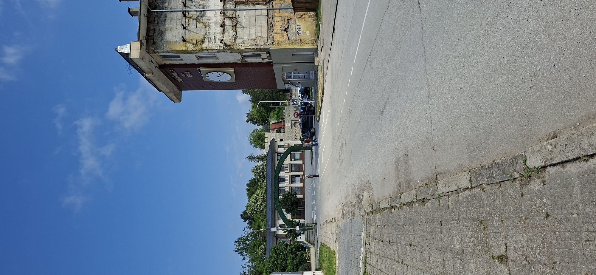 This is an outdoor street scene captured in Strazhitsa, Bulgaria, during bright daylight under a clear blue sky with scattered white clouds.
The image depicts a street with an asphalt road and a concrete sidewalk to its right. On the left side of the street stands a large, multi-story building with a weathered facade, showing signs of age with cracked plaster and exposed bricks. A prominent feature of this building is a large clock face centrally placed on a brown section. Above the clock, text is visible, reading "ВИНОПРОИЗВОДСТВО" (Winemaking/Wine Production) and below it, the year "1966". Further up on the weathered yellow wall of the same building, the year "1948" is painted.
Further down the street, on the left, another multi-story building with a more contemporary appearance is visible, featuring numerous windows. In front of this building, a distinctive green archway, possibly a decorative entrance or monument, spans part of the view.
Several people are present in the scene; a few individuals are standing or walking on the sidewalk near the green archway and further down the street. Several cars are parked or moving in the distance along the street on the right. Lush green trees are abundant, especially in the background and surrounding the buildings, indicating a green environment. A red "STOP" sign is partially visible on the right side of the street. The time of day appears to be midday or early afternoon given the bright lighting and minimal shadows.