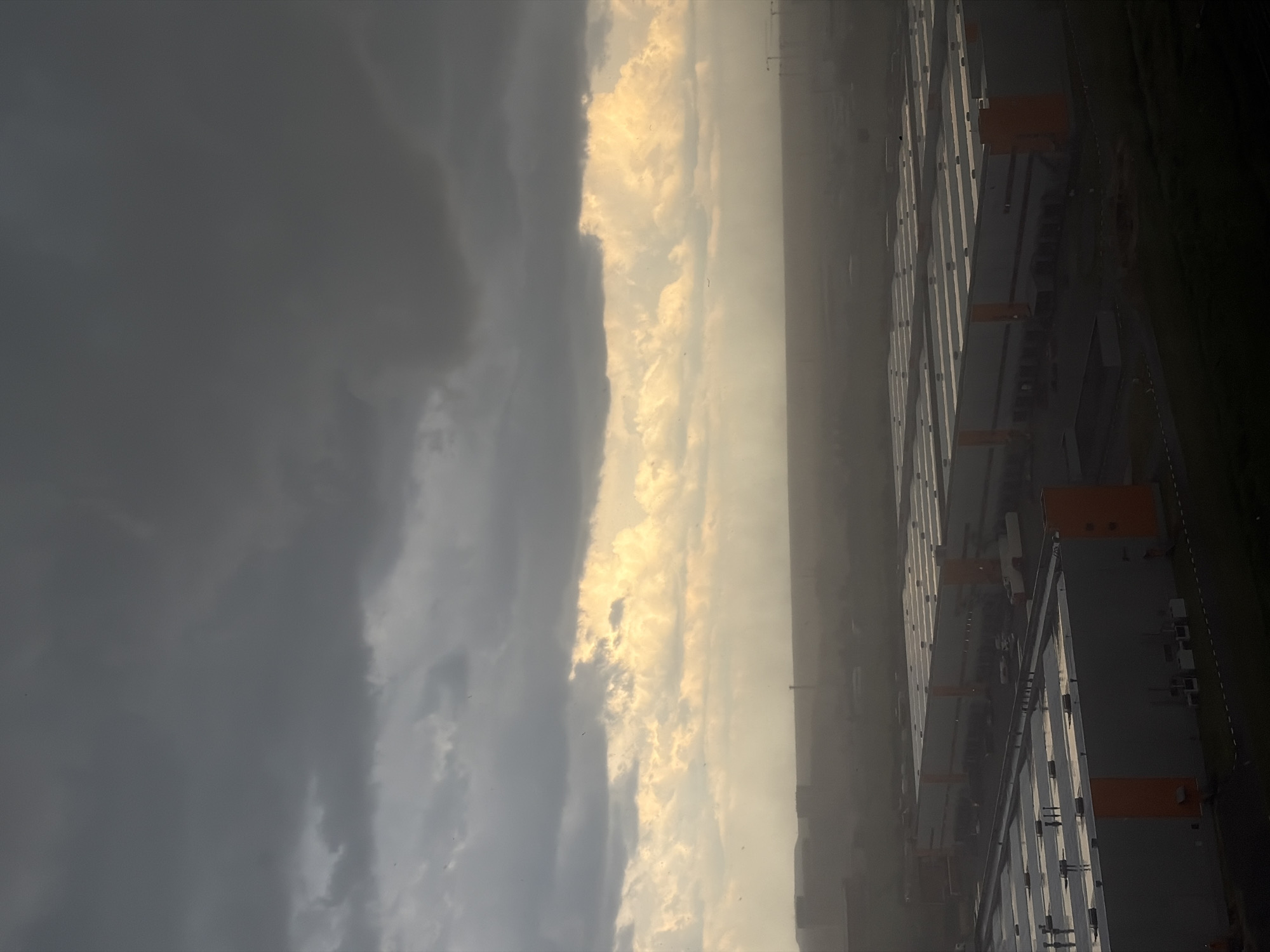 The image depicts a vast industrial area under a dramatic sky. In the foreground, rows of large, modern warehouses with grey facades and orange accents are visible. Their flat roofs are illuminated by a diffuse light, suggesting overcast conditions. The warehouses are arranged in a grid pattern, indicative of a planned industrial zone. A dark, misty haze hangs over the middle ground, obscuring the details of more distant structures and a faint silhouette of what appears to be a cityscape or more industrial buildings. The sky is dominated by heavy, dark grey storm clouds, with a bright, yellowish band of light breaking through in the middle distance, hinting at the sun's position behind the clouds. A few specks, possibly birds or dust particles, are visible against the sky. The overall atmosphere is one of brooding weather over an expansive industrial landscape. This scene is consistent with the industrial zones found in the outskirts of Saint Petersburg, Russia, such as the Kolpino district or the areas surrounding the Pulkovo Airport, where large logistics and manufacturing facilities are common.