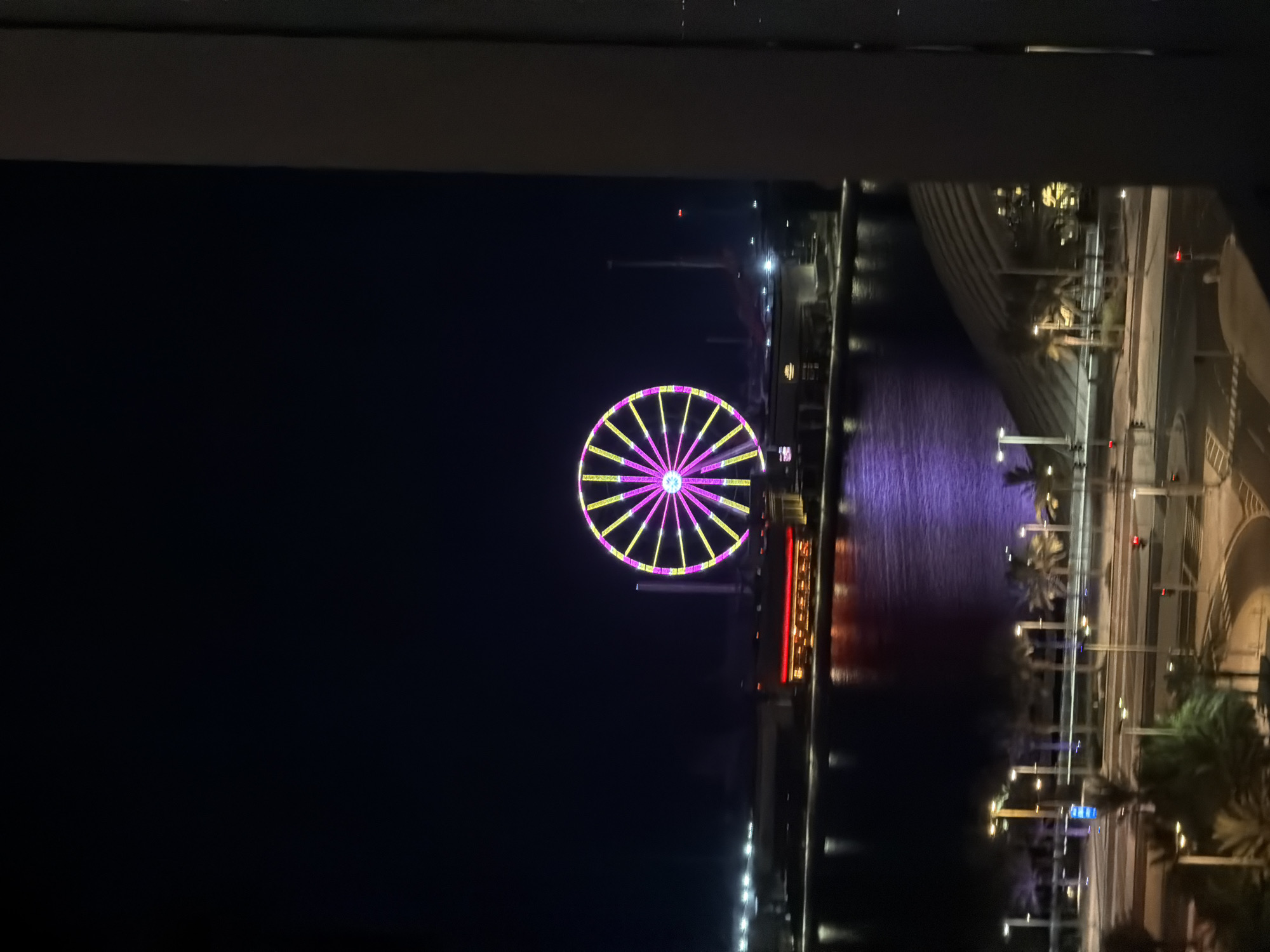 The image shows a nighttime scene in a city. A large Ferris wheel, illuminated with pink and yellow lights, is the central focal point. It is situated across a body of water, which reflects the lights of the city and the Ferris wheel. In the foreground, there is a road with streetlights and palm trees, suggesting a warm climate. The overall impression is that of a vibrant urban entertainment district at night.
