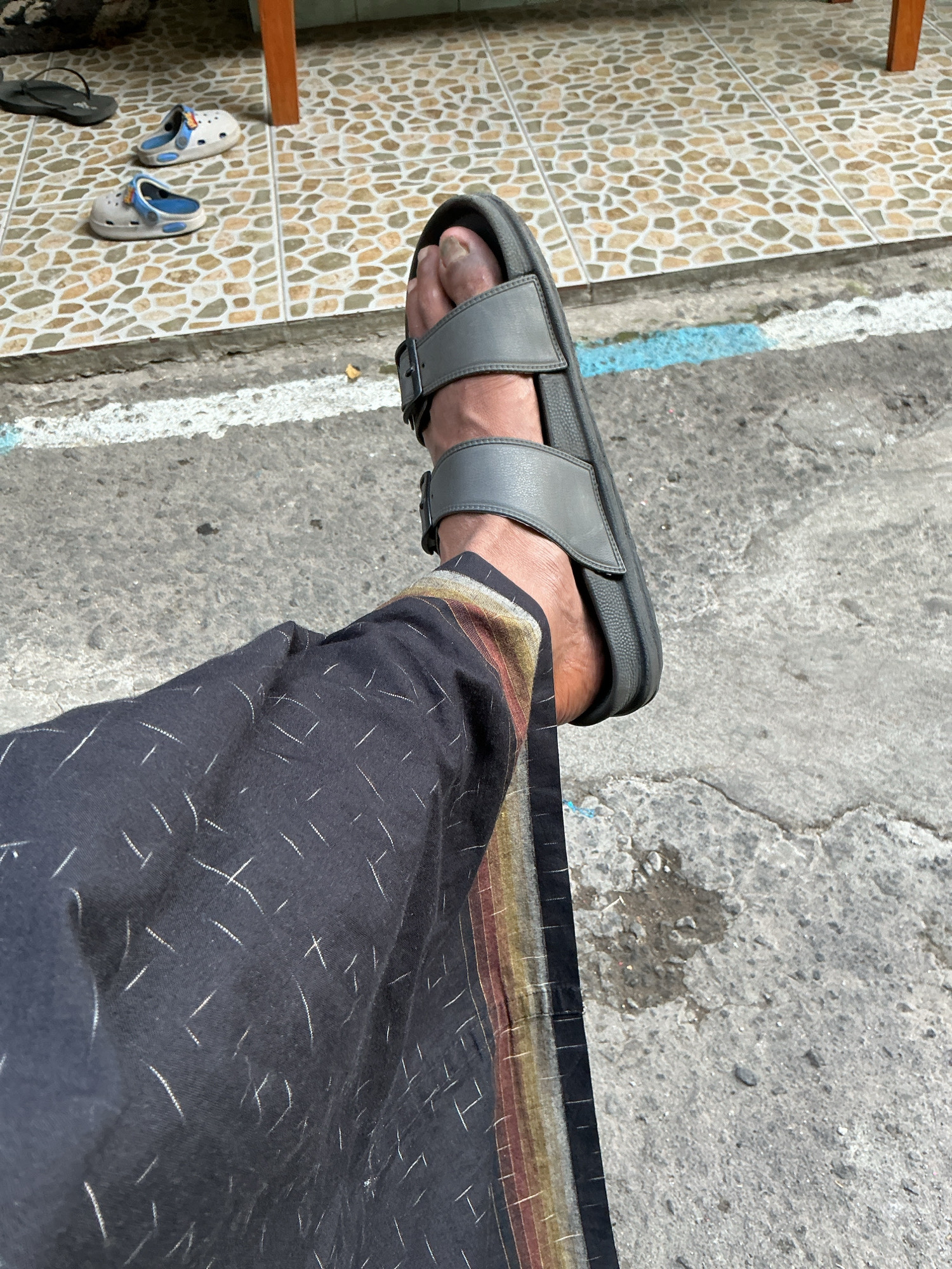 In Magelang, Indonesia, a person's leg is shown from the knee down, resting on a concrete sidewalk. The person is wearing dark pants with a white, geometric pattern and a border of thin, vertical stripes in red, yellow, and brown. On the person's right foot, they are wearing a gray sandal with a single buckle strap. In the background, there are tiled floors and several pairs of children's sandals, including white and blue Crocs and black flip-flops. The overall scene appears to be an outdoor area, possibly a residential or commercial space.