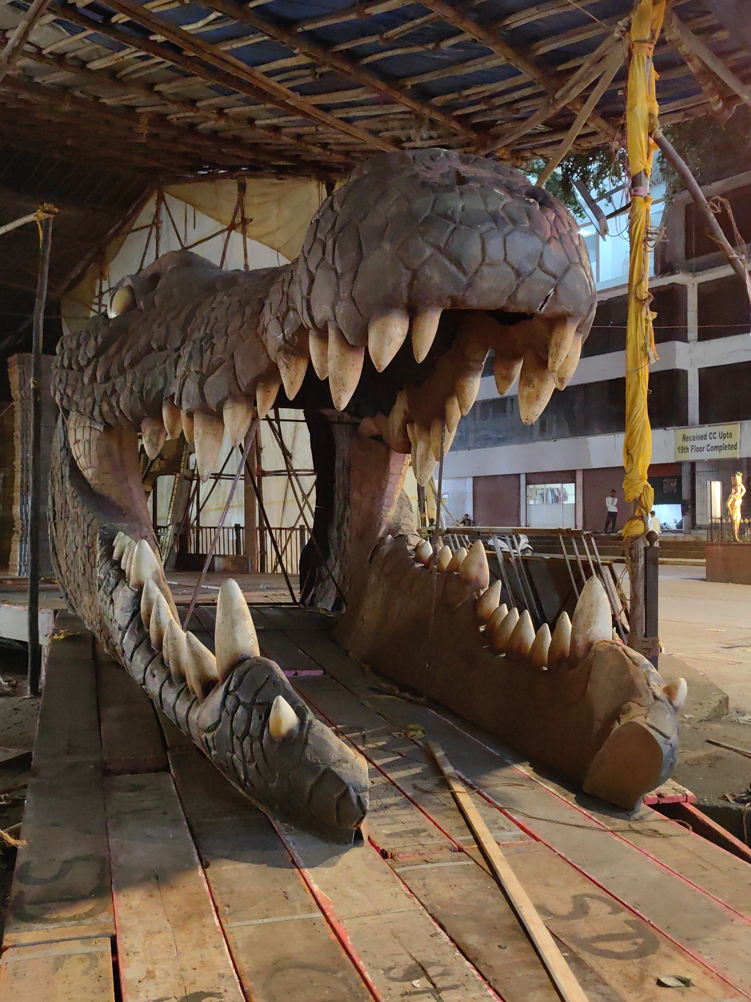 The image shows a large, detailed sculpture of a crocodile's head and open jaws, seemingly under construction or a temporary installation. The sculpture is made of textured materials, possibly foam or fiberglass, painted in earthy tones to resemble scales. Its teeth are large, sharp, and conical. The crocodile is positioned on a wooden platform, with scaffolding and construction materials visible in the background. The setting appears to be outdoors, possibly an event preparation area or a construction site in a city.
In the background, a building with multiple floors is visible, with signs indicating "Received CC Upto 19th Floor Completed." Several people are present in the background, some appearing to be workers or onlookers. The overall atmosphere suggests an urban environment in Mumbai, India, likely preparing for a festival or public event. The lighting suggests it may be late afternoon or early evening.