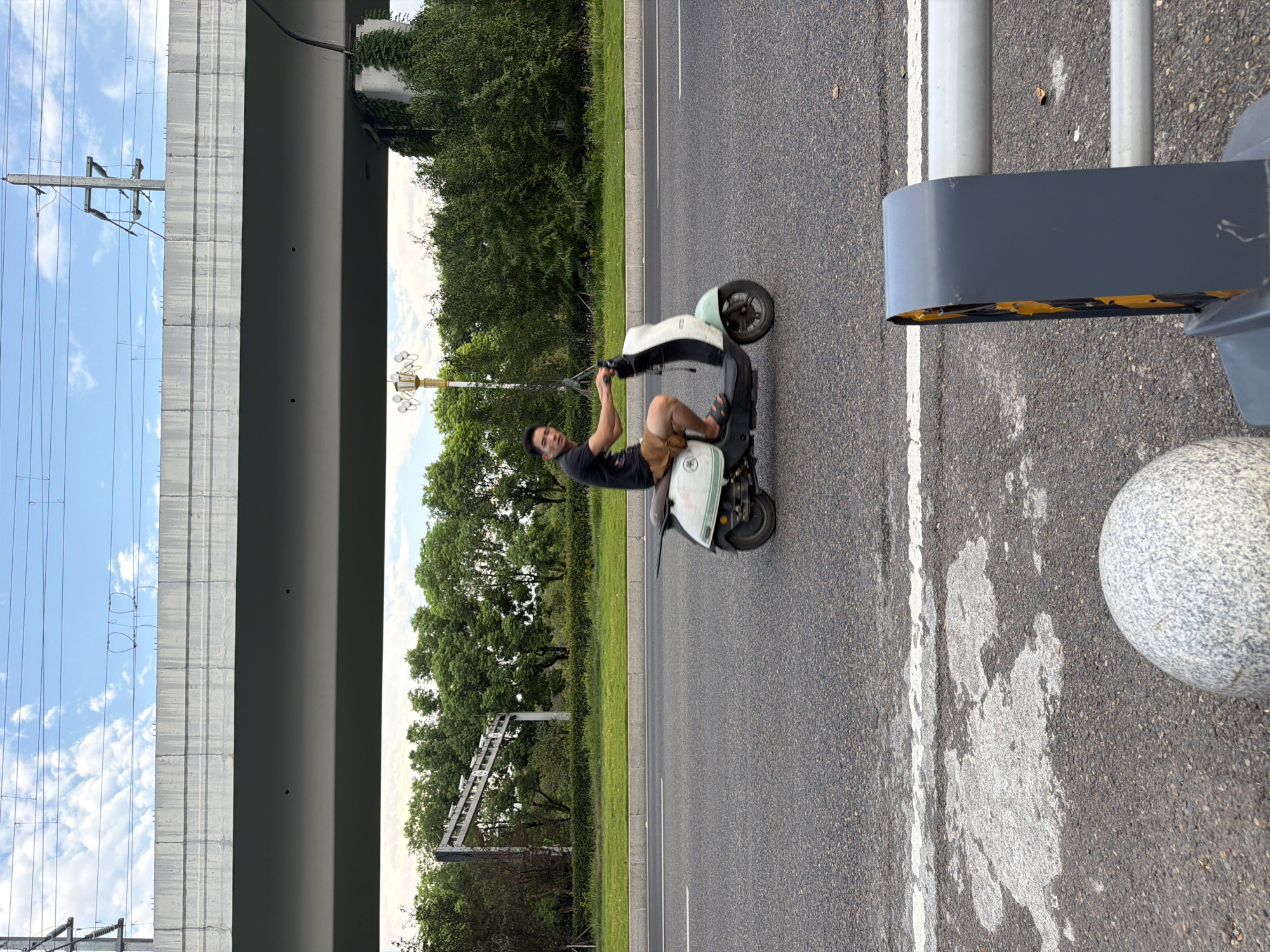 An Asian male, appearing to be in his late 20s to early 30s, rides a light-colored electric scooter on an asphalt road. He is wearing a black t-shirt and brown shorts. The scooter is white and light green. The background features a raised concrete overpass, trees, a blue sky with clouds, and power lines. In the foreground, there is a light gray sphere and a metal barrier. The scene is located in the city of Jiangjin, China, as indicated by the context provided.